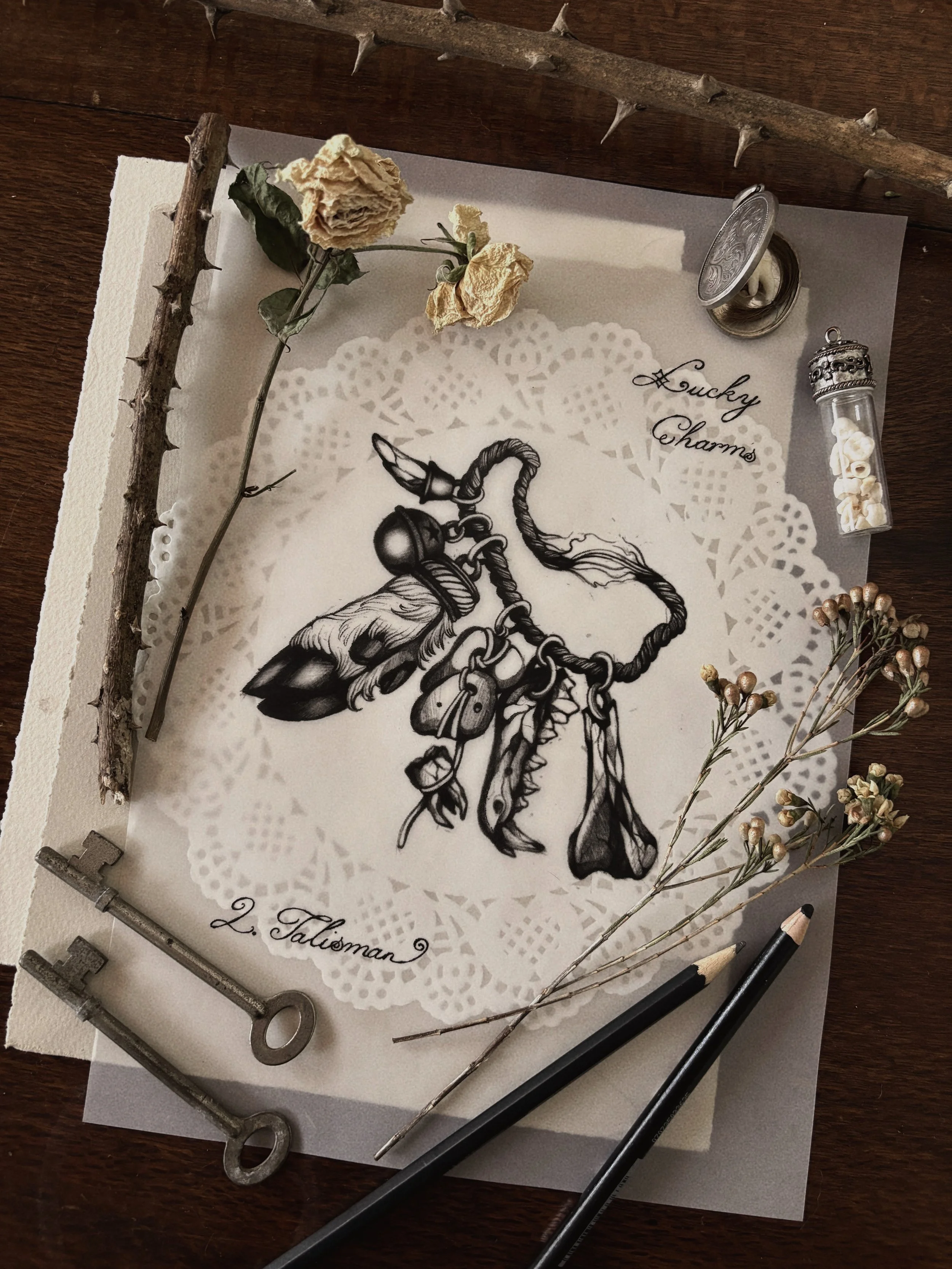 A black-and-white drawing of a chain with a bell on it, surrounded by dried flowers and vintage keys, on a decorative paper with the words 'Lucky Charms' and '2. Talisman'.