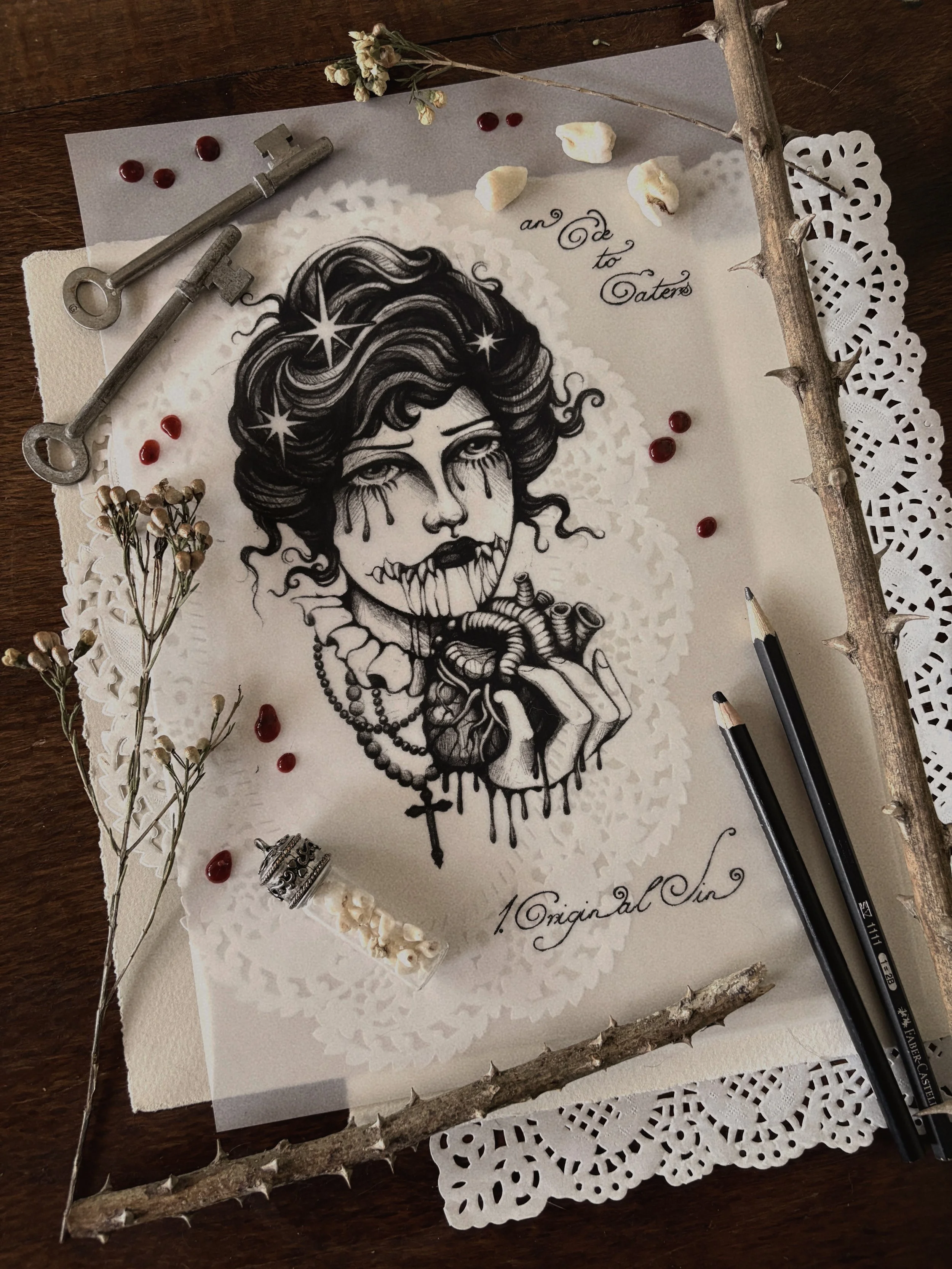 A black and white illustration of a woman with makeup and jewelry, with the words 'an ode to Eaters' and 'Original Sin'. The artwork is surrounded by decorative items, including dried flowers, a twig, a piece of lace, two black pens, two old keys, a small crown, and scattered red and white beads, all on a wooden surface.