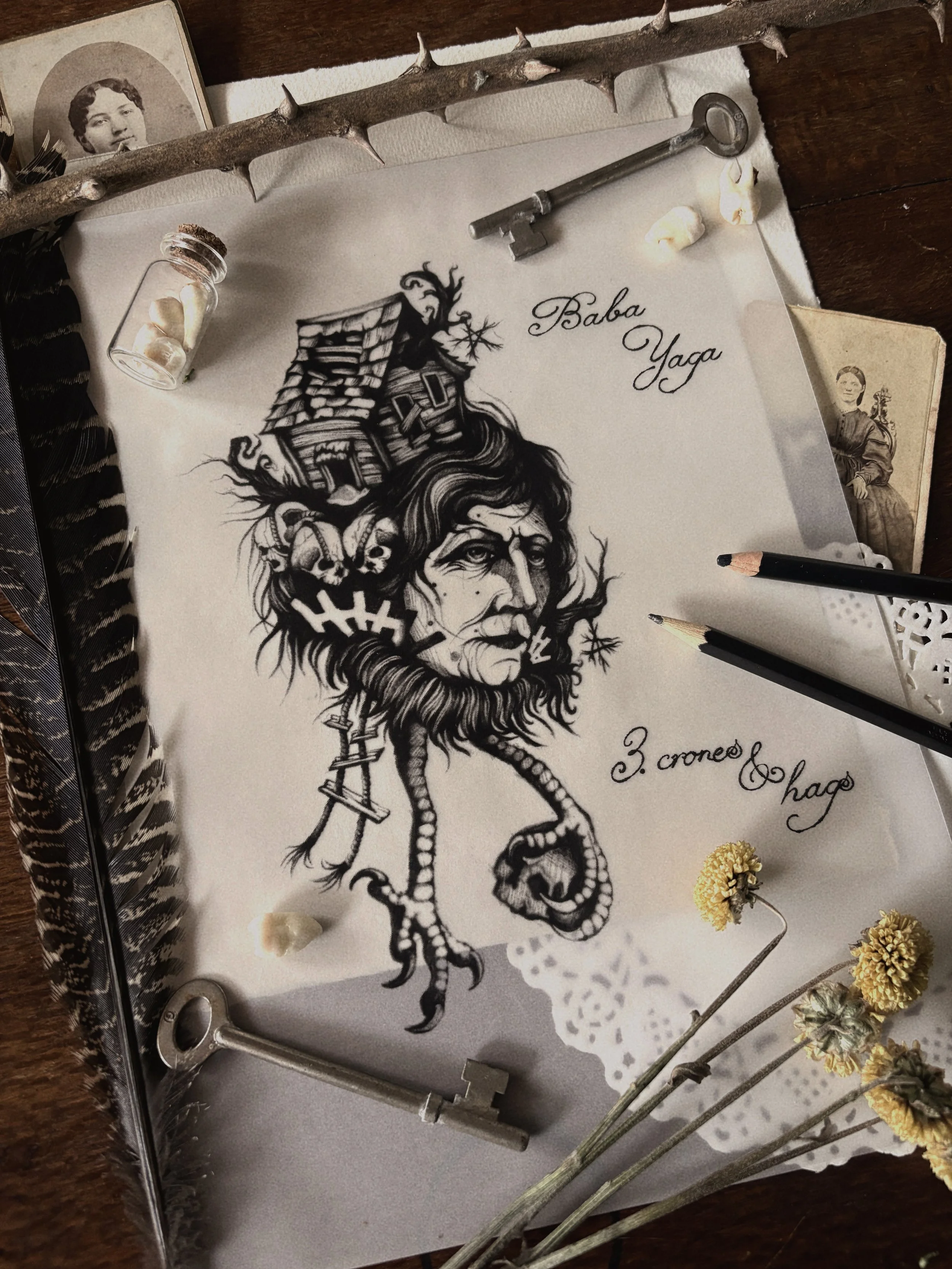 A detailed black and white drawing of a mythological creature with the head of an old woman and a body of a serpent, with a house on top of her head. The drawing is on a piece of paper with the words "Baba Yaga" written on it, and some other text and decoration. Surrounding the drawing are dried flowers, keys, old photographs, and various papers.