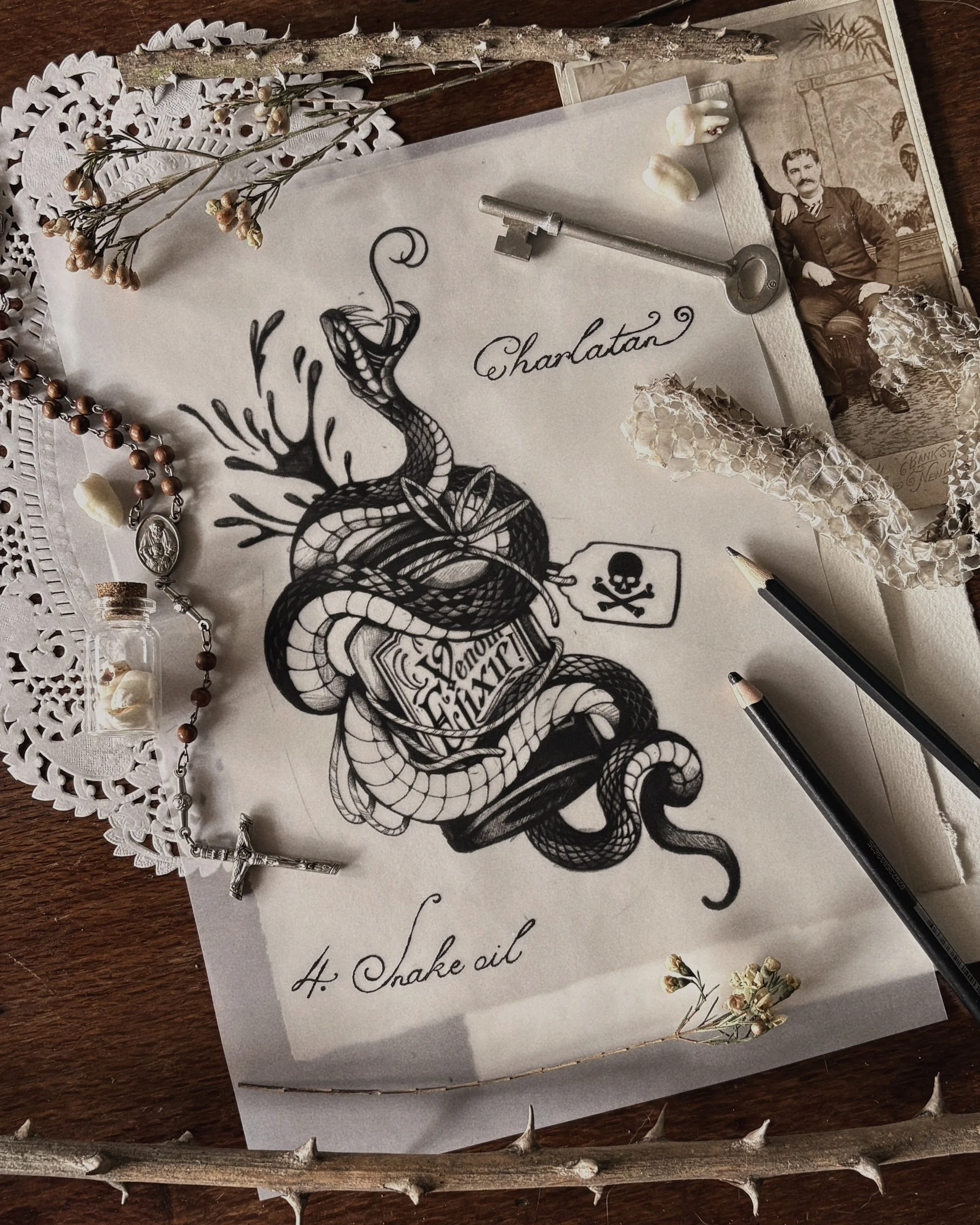 A vintage sketch of a snake wrapped around a jar labeled 'Wake oil' is on top of a handwritten note that says 'Charlotte'. The scene includes dried flowers, a key, a beaded necklace, a small jar, a twig, and a vintage photograph of a man, all arranged on a wooden surface with a decorative doily and a large thorny branch surrounding the items.