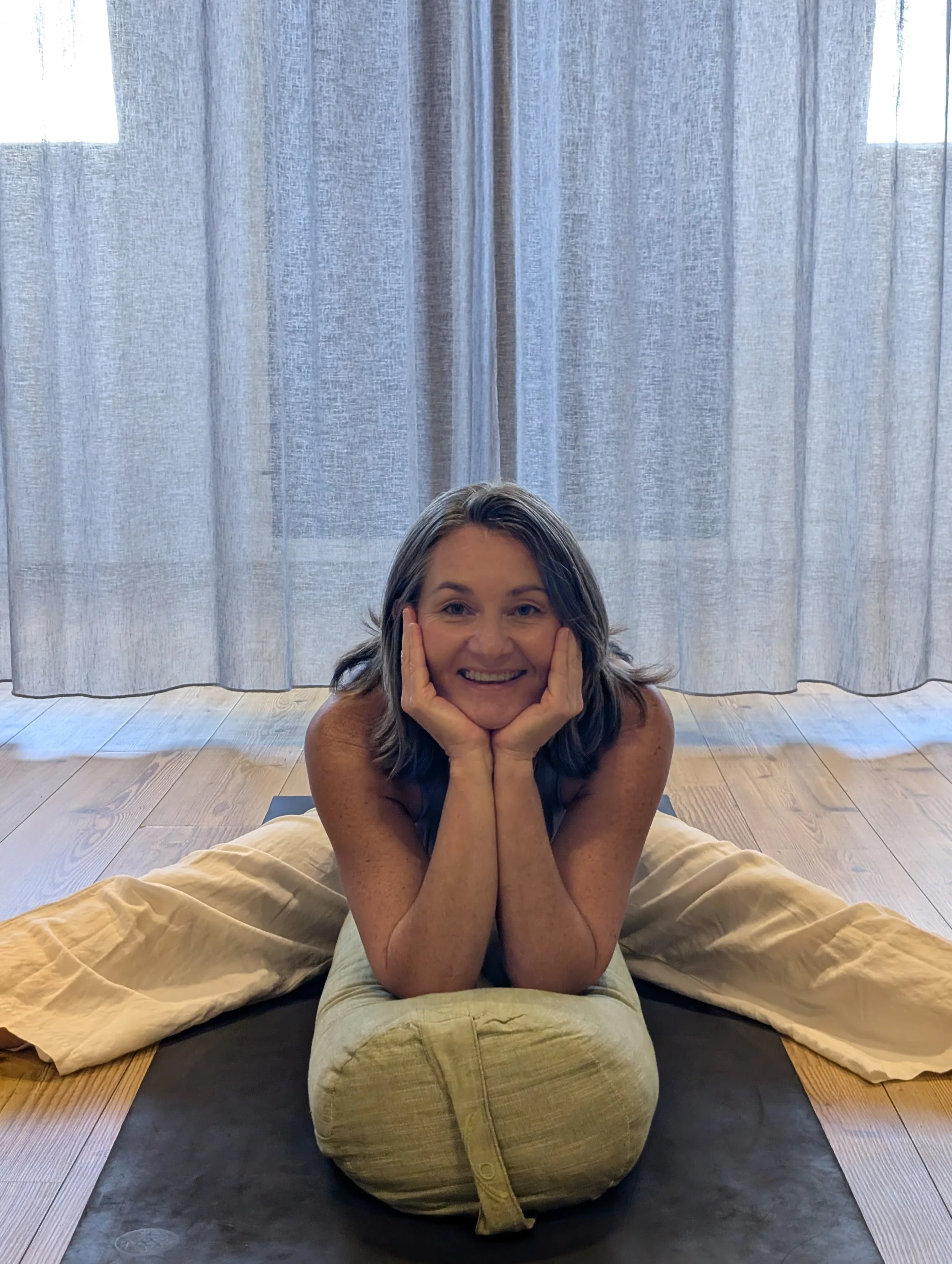 A woman practicing yoga on a mat in a room with curtains and wooden floors, smiling and resting her face in her hands while lying on her stomach.