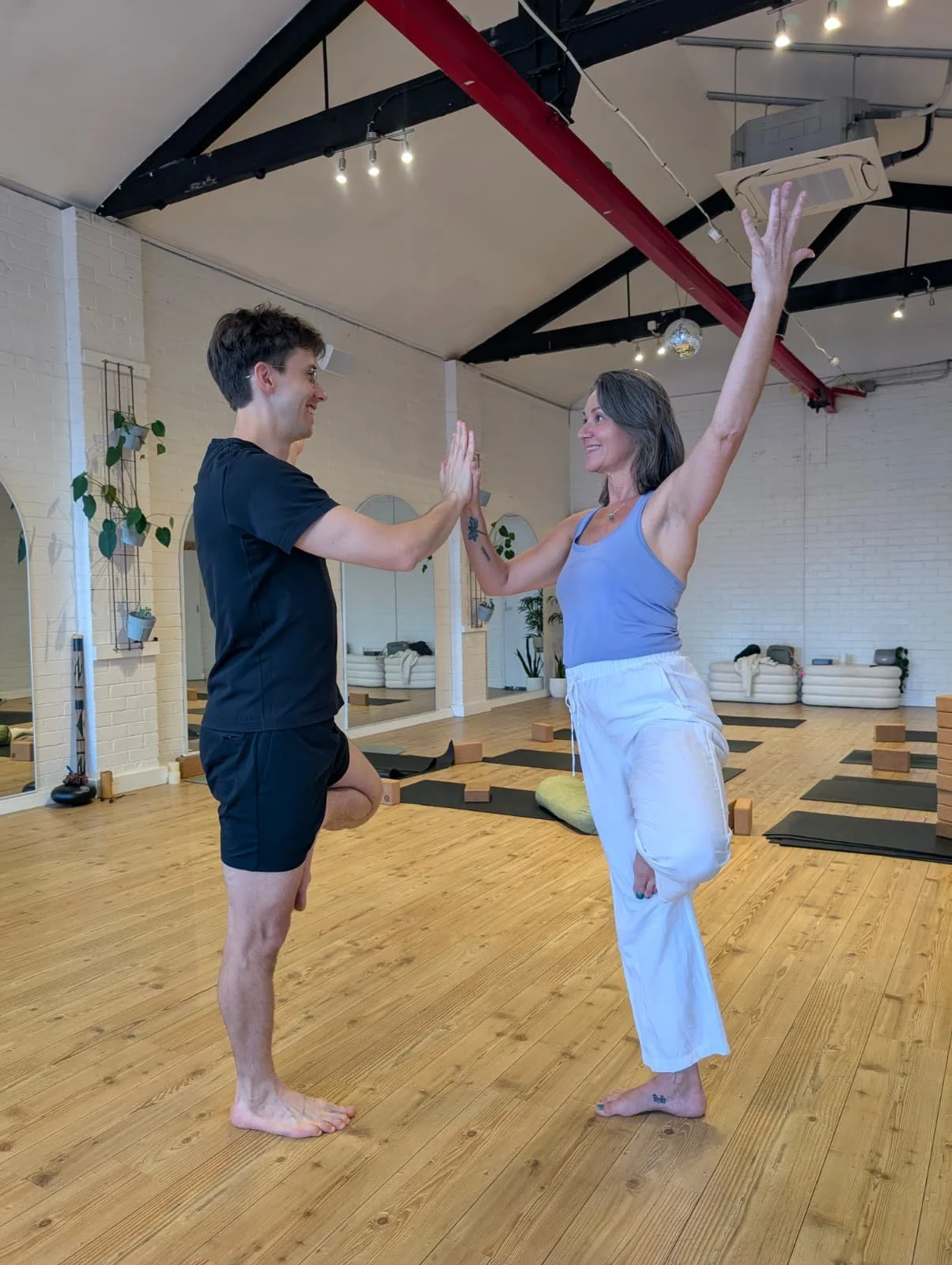 Two people practicing yoga in a studio, facing each other with high fives, one standing on one leg and the other raising an arm.