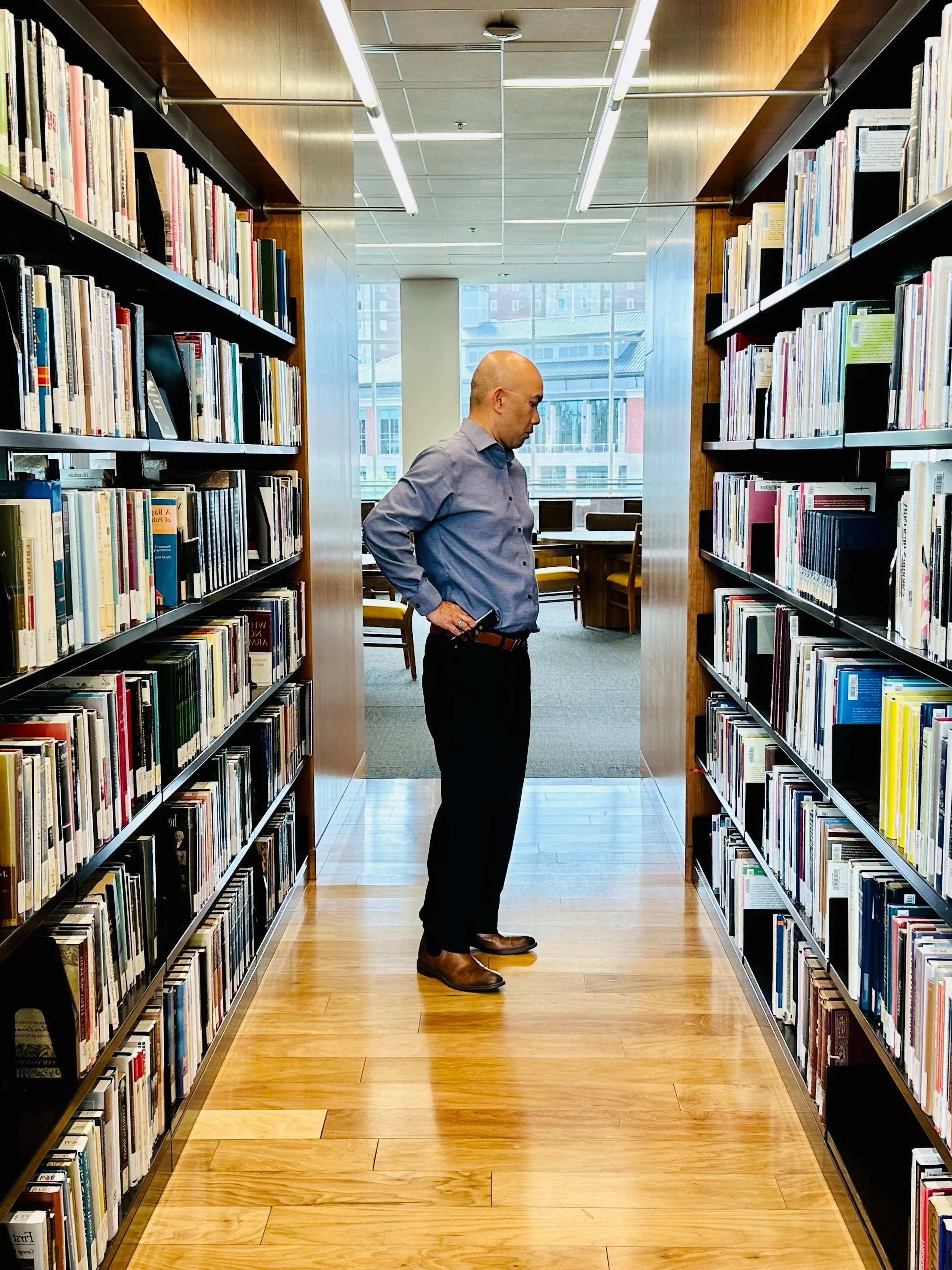 A man with a shaved head wearing a gray shirt and black pants standing in a library aisle between bookshelves, looking at books. He is holding a phone in his right hand behind his back. The library has wooden floors, shelves filled with books, and large windows with a city view in the background.