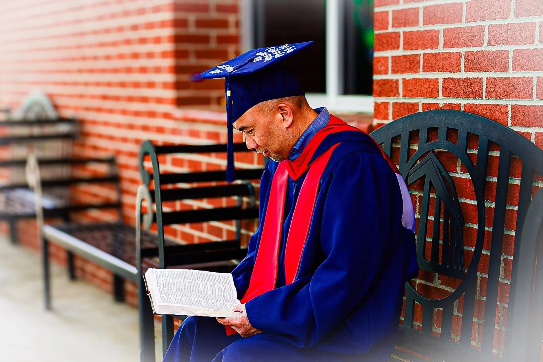 A man in a blue graduation gown and cap with a red stole sitting on a black metal bench, reading a book outside against a red brick wall.