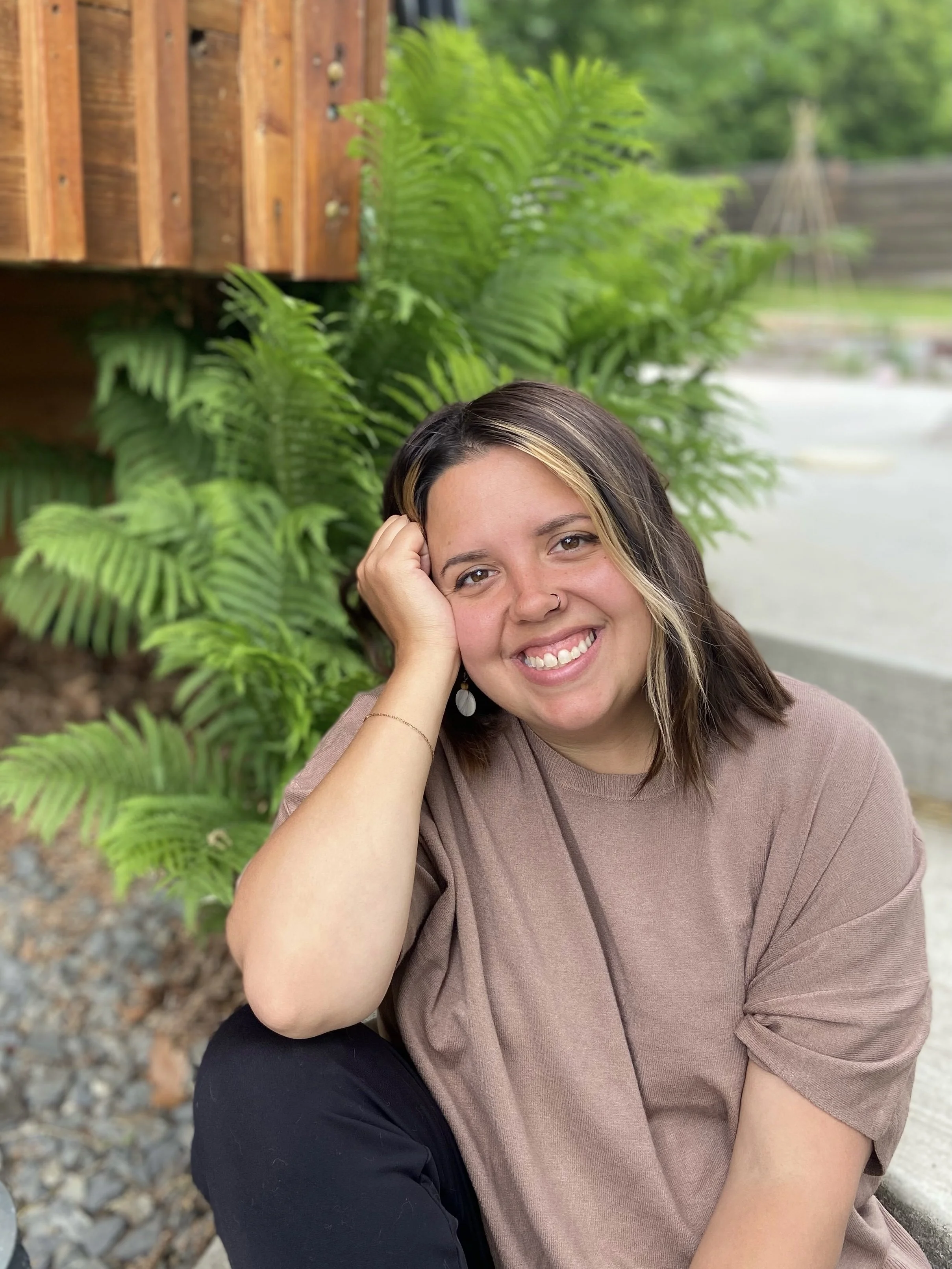 A smiling woman with dark hair and blonde highlights, sitting outdoors near green plants, with a wooden fence in the background.