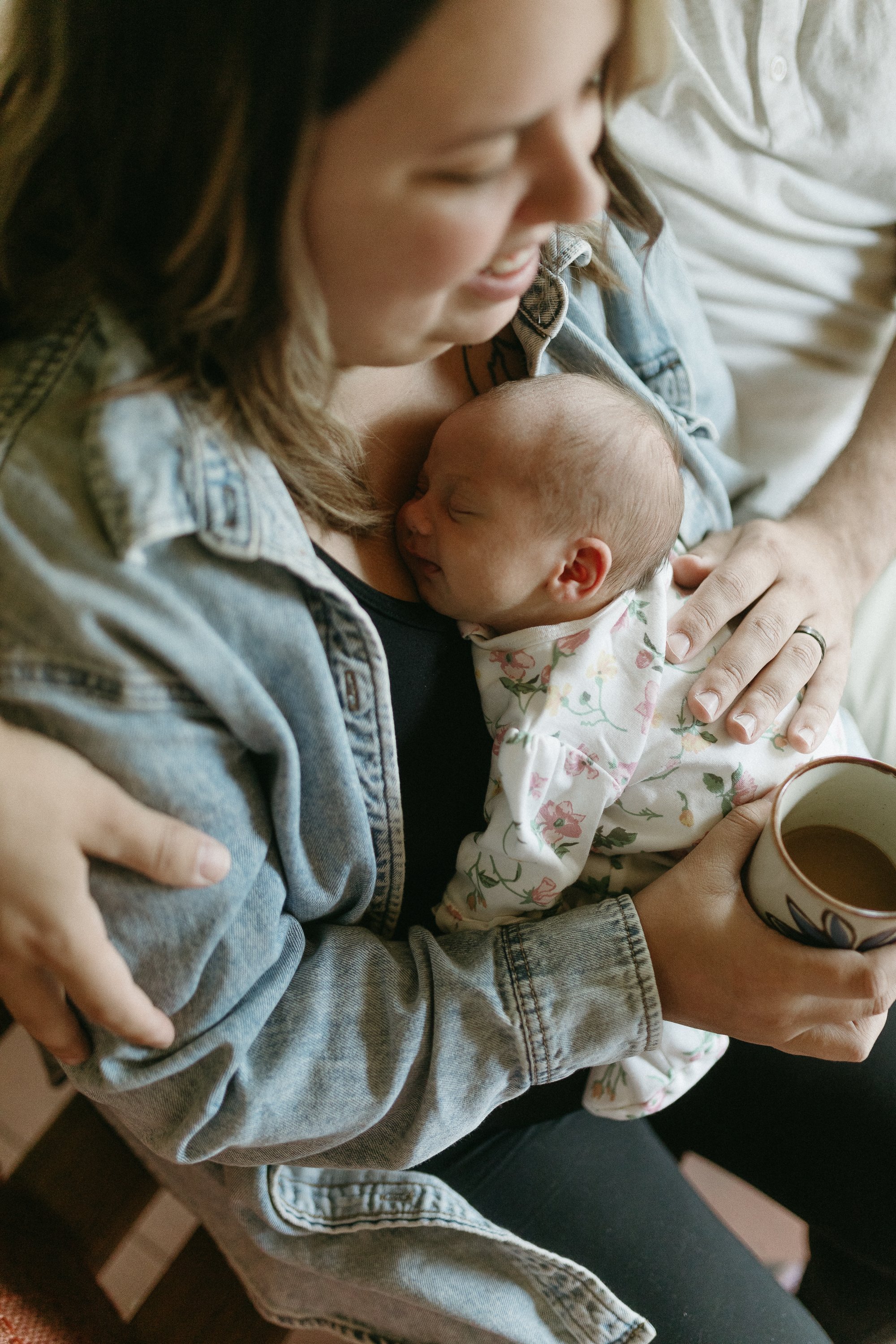 A woman holds a sleeping baby while sitting on a person, holding a cup of coffee in her hand.