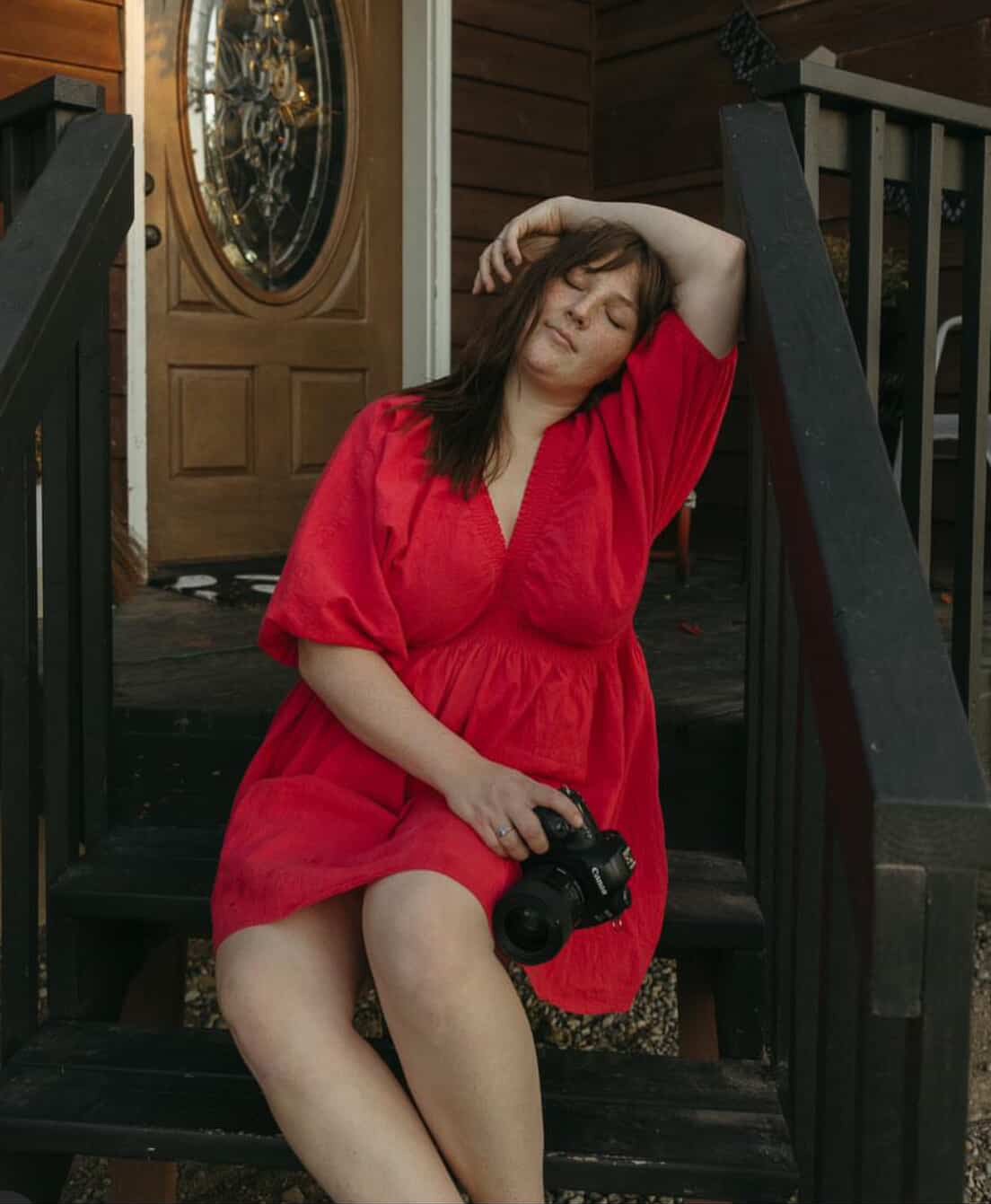 A woman in a red dress sitting on the front steps of a house, holding a camera in her lap, with one arm raised and her eyes closed.