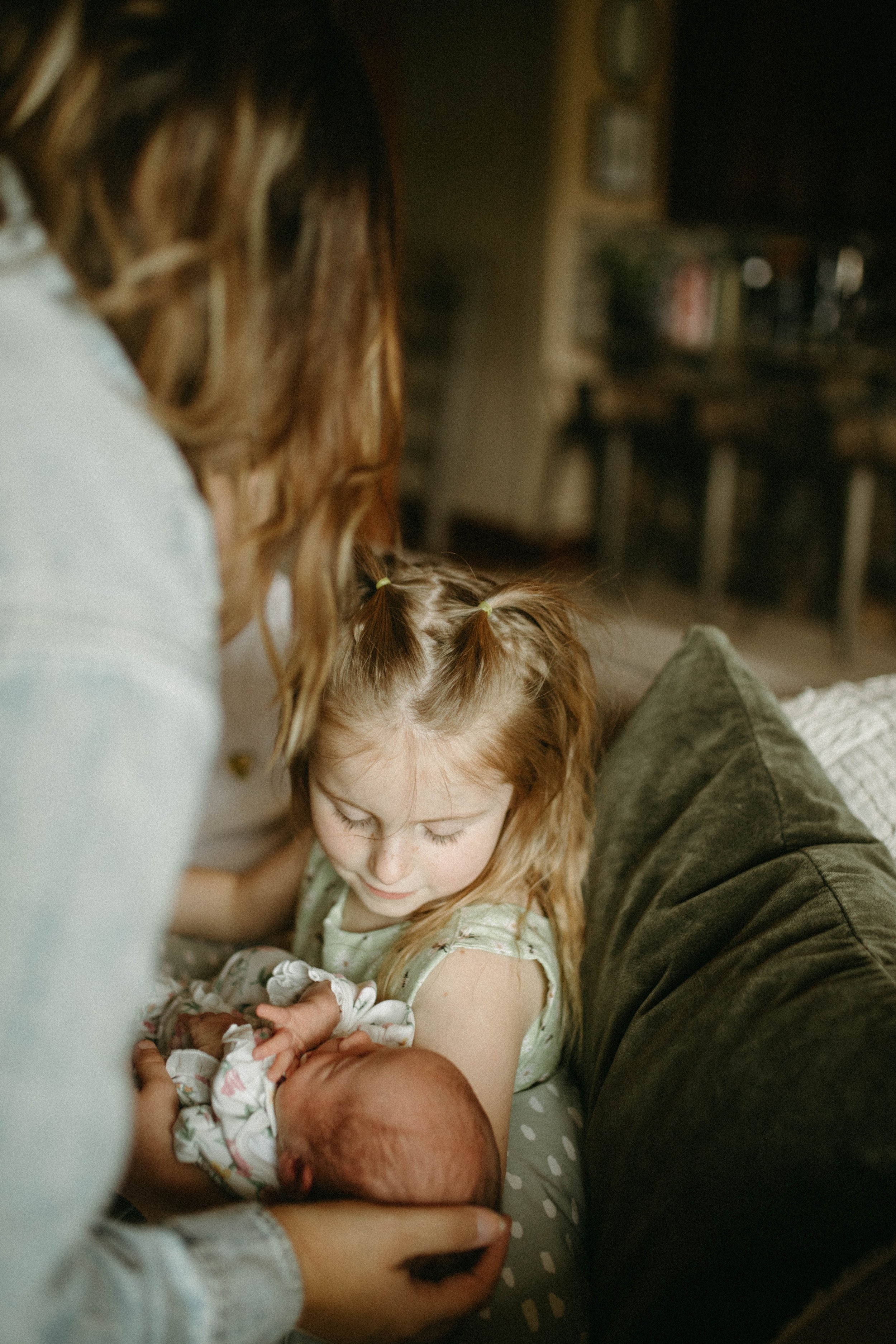 A young girl with red hair, sitting on a sofa, looking down at a newborn baby she is holding, with a woman supporting the baby. The scene appears warm and tender, set in a cozy living room.