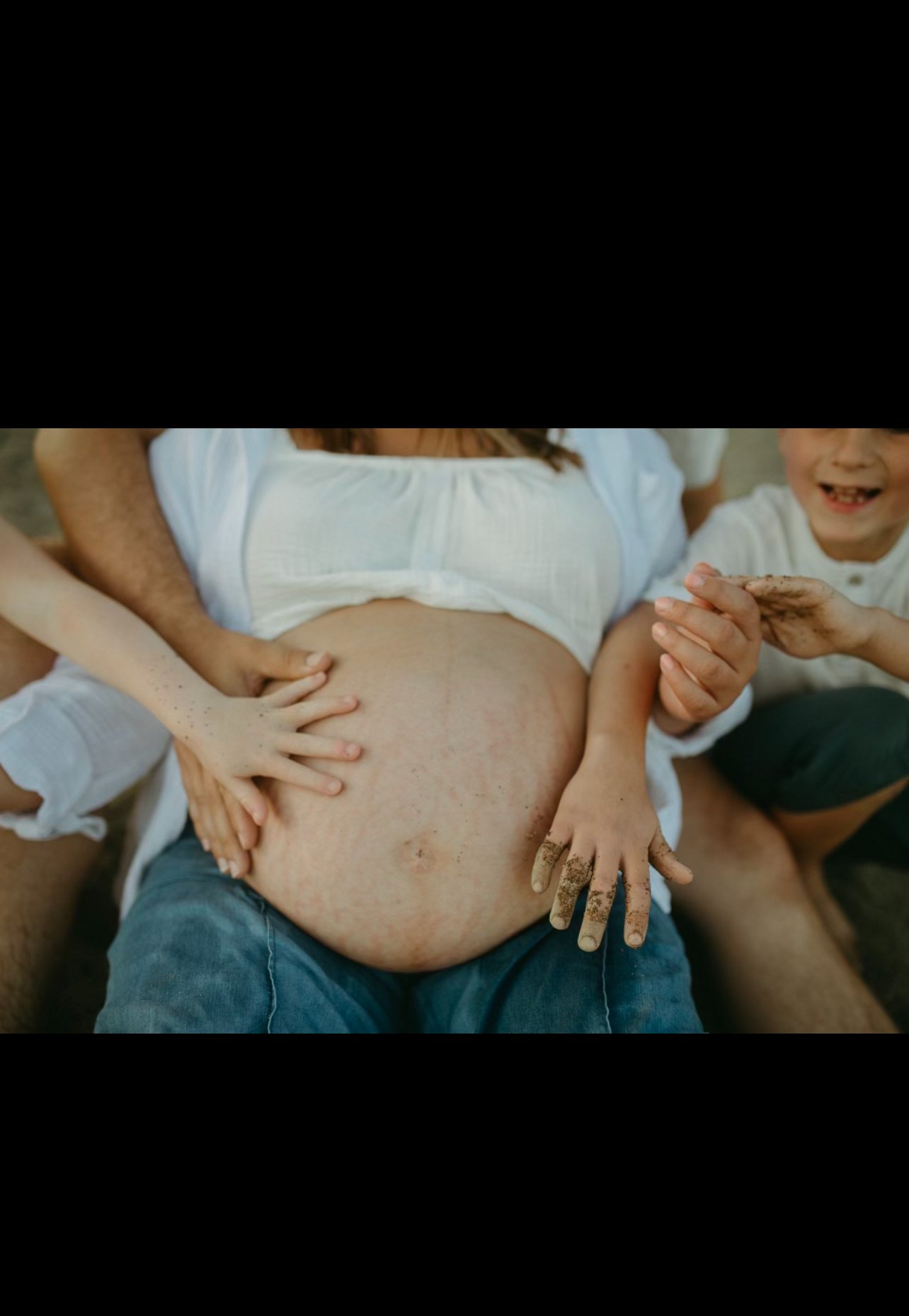 Close-up of a pregnant woman with children touching her belly, smiling, and playful with muddy hands, in a joyful outdoor scene.
