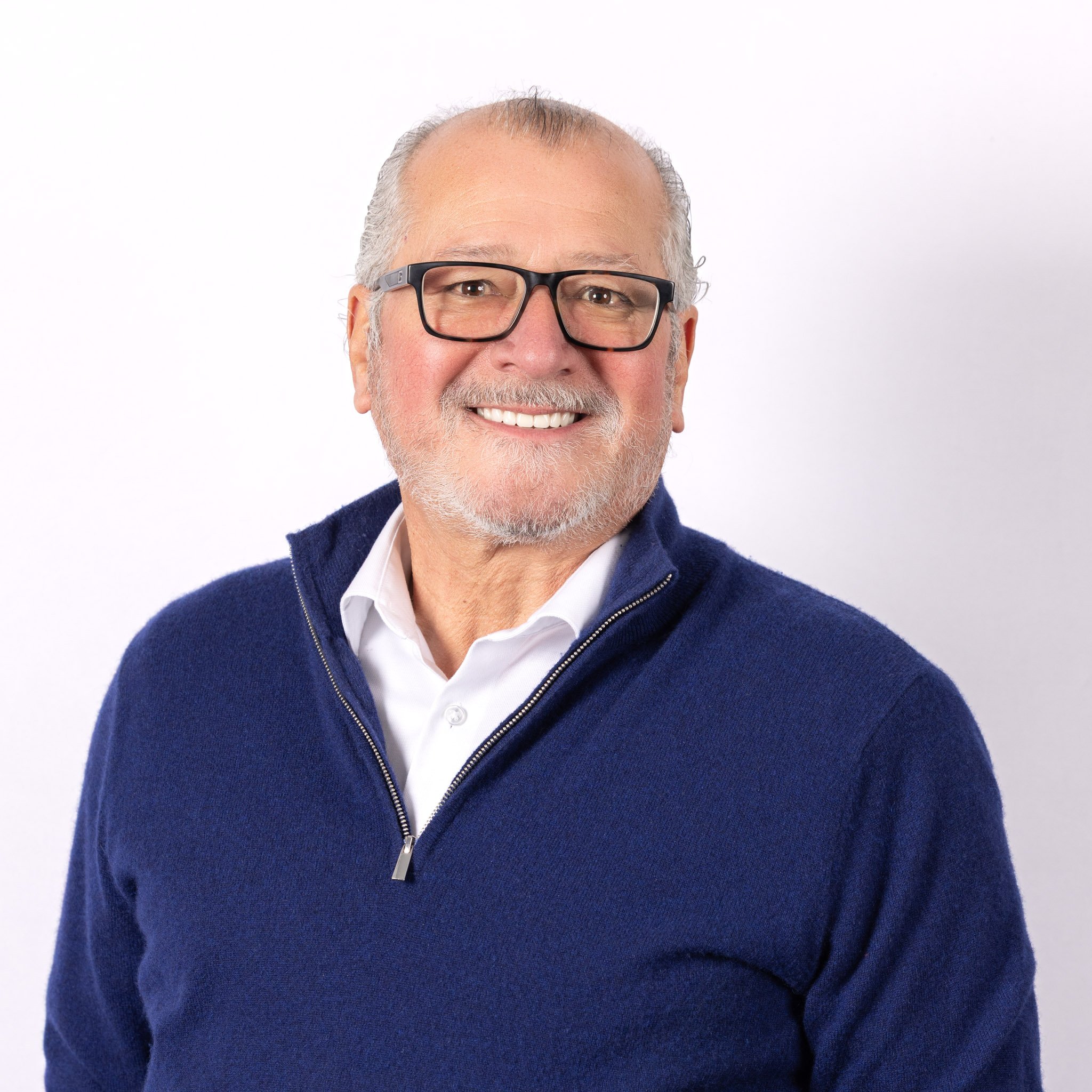 Smiling older man with gray hair, beard, and glasses wearing a white shirt and navy blue zip-up sweater against a plain white background.
