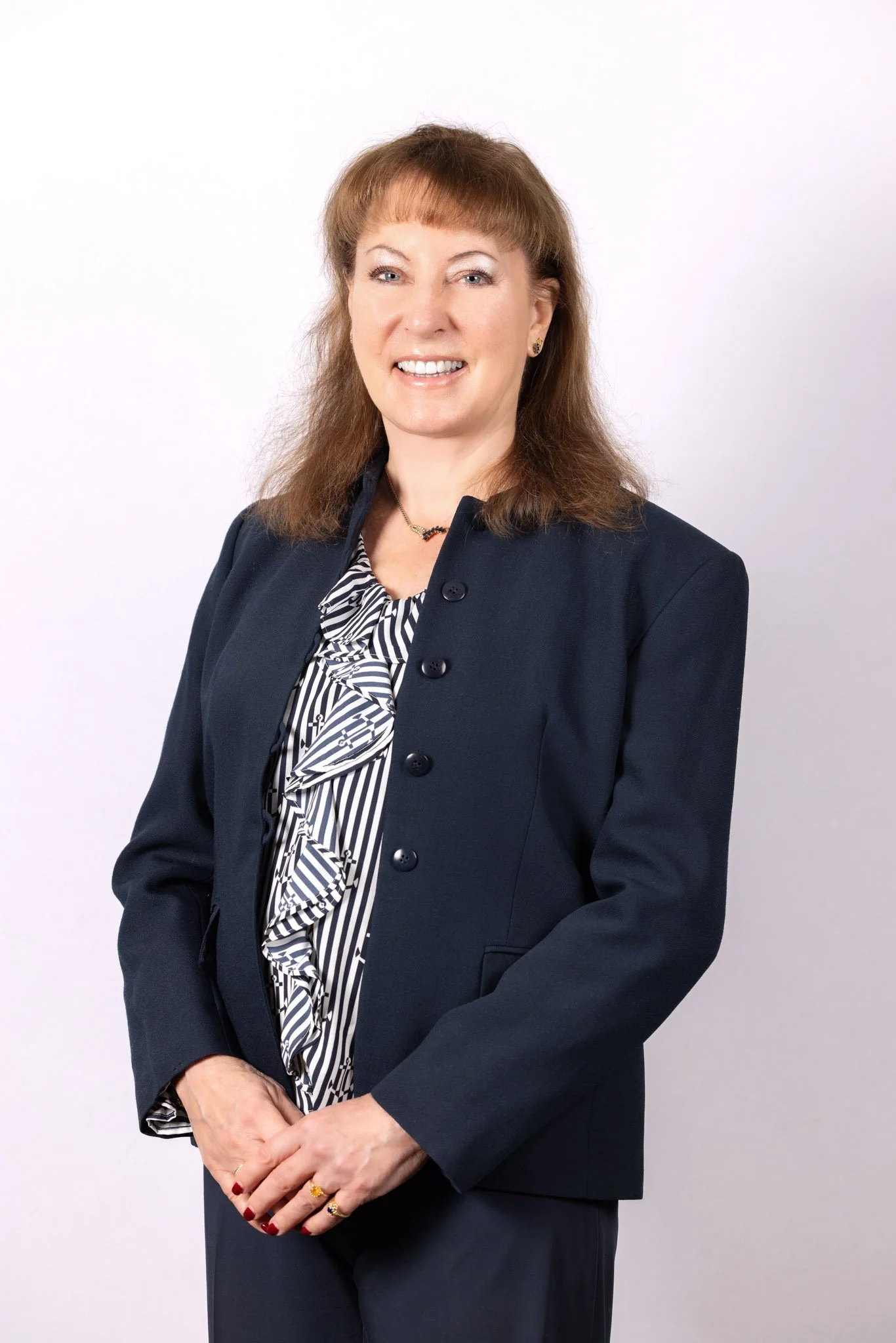 A professional woman with reddish-brown hair wearing a navy blazer over a black and white ruffled striped blouse, standing against a light background, smiling.