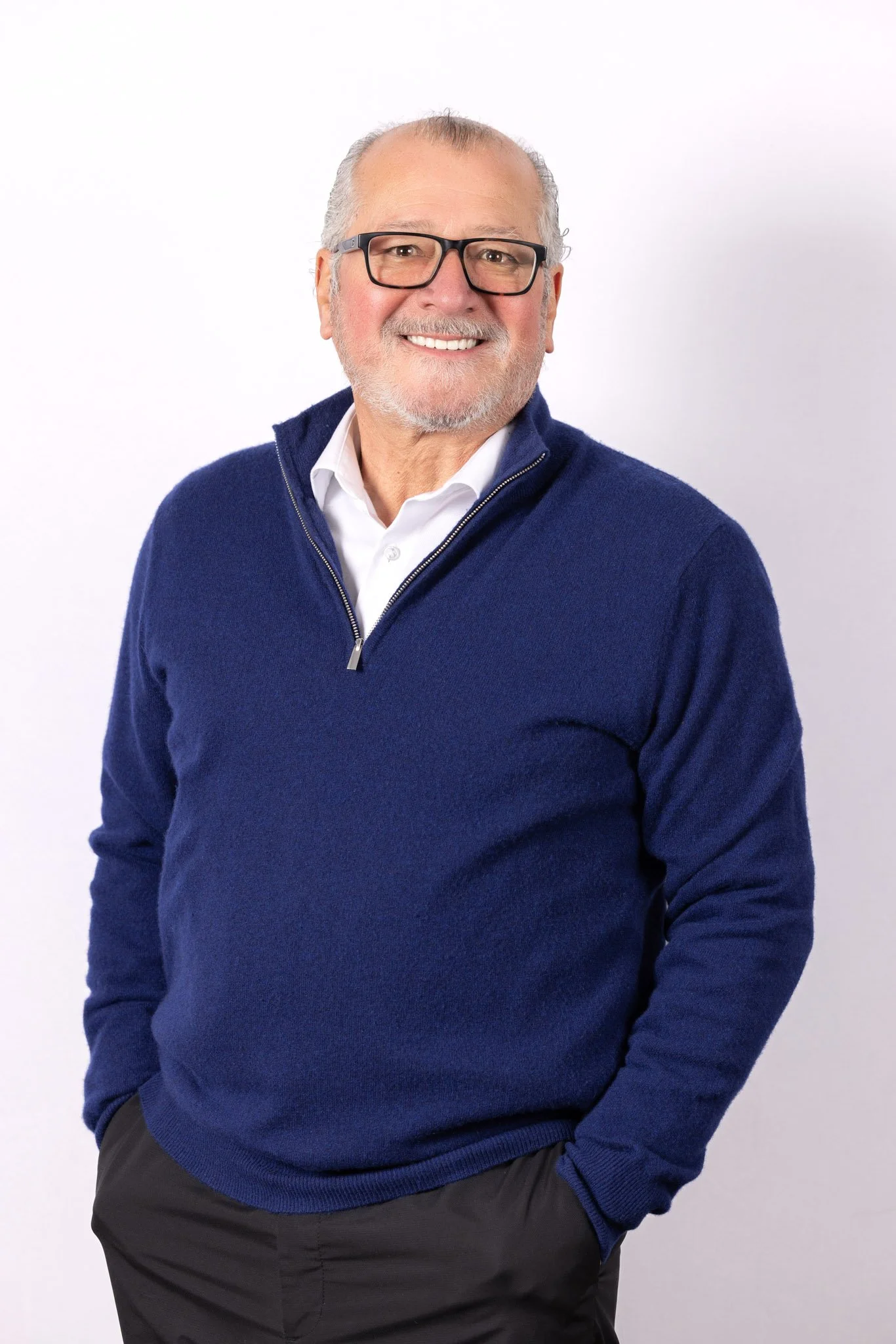 A smiling older man with gray hair, wearing glasses, a white shirt, and a blue zip-up sweater, posing against a plain white background.
