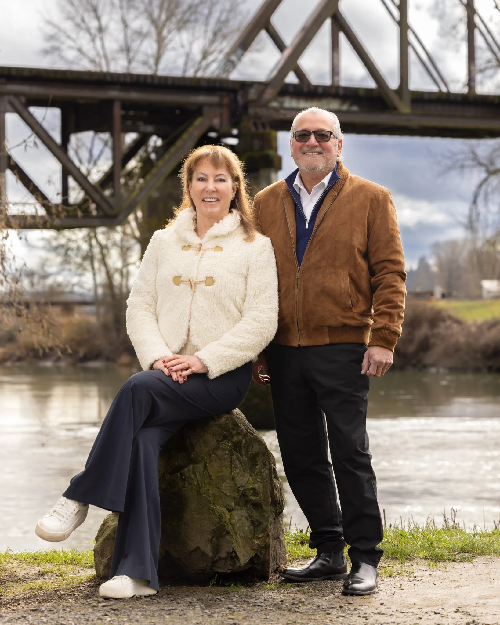 A smiling middle-aged woman in a cream-colored jacket and navy pants sitting on a rock, alongside a middle-aged man in a brown jacket and black pants standing by her side, with a river and an old wooden bridge in the background.
