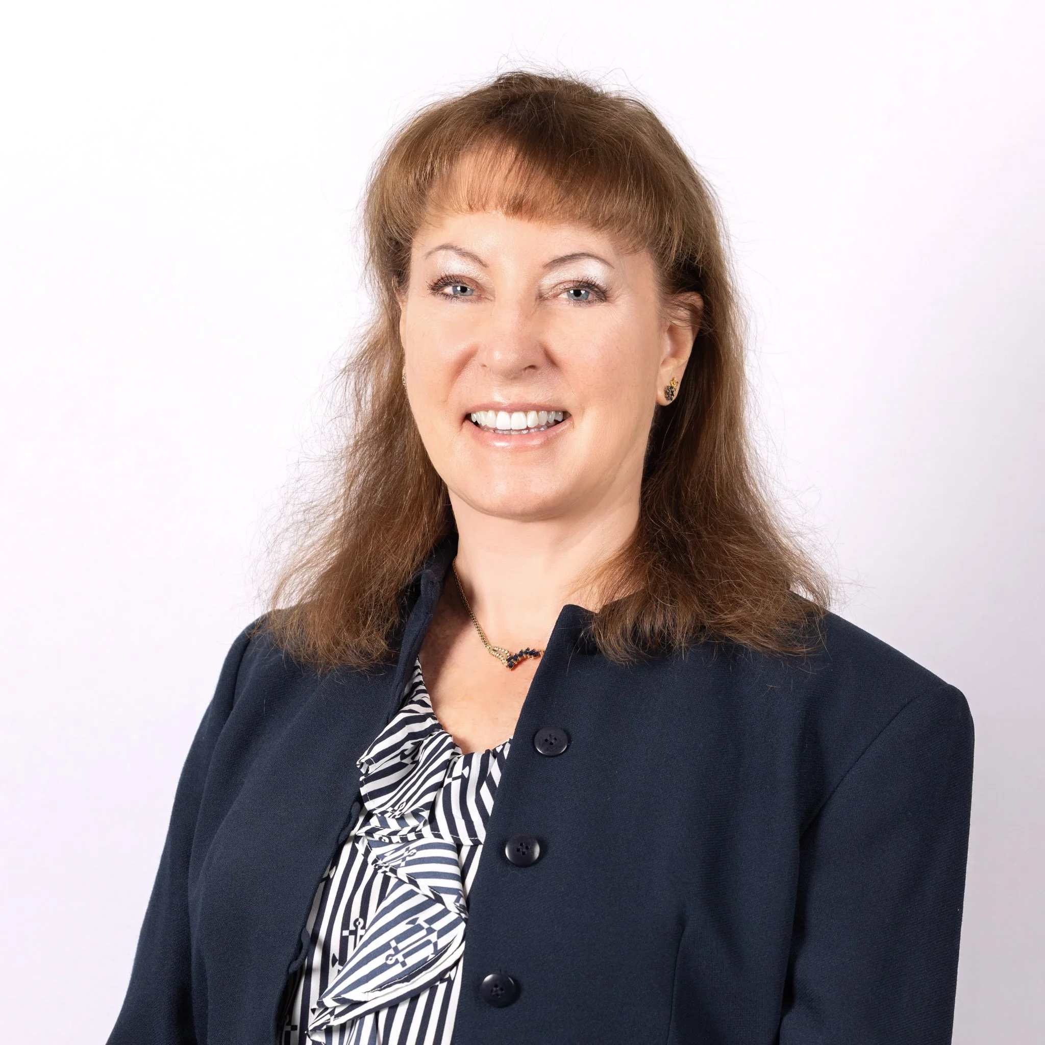 A professional woman with shoulder-length brown hair, smiling, wearing a dark blazer, patterned blouse, and jewelry, standing against a white background.