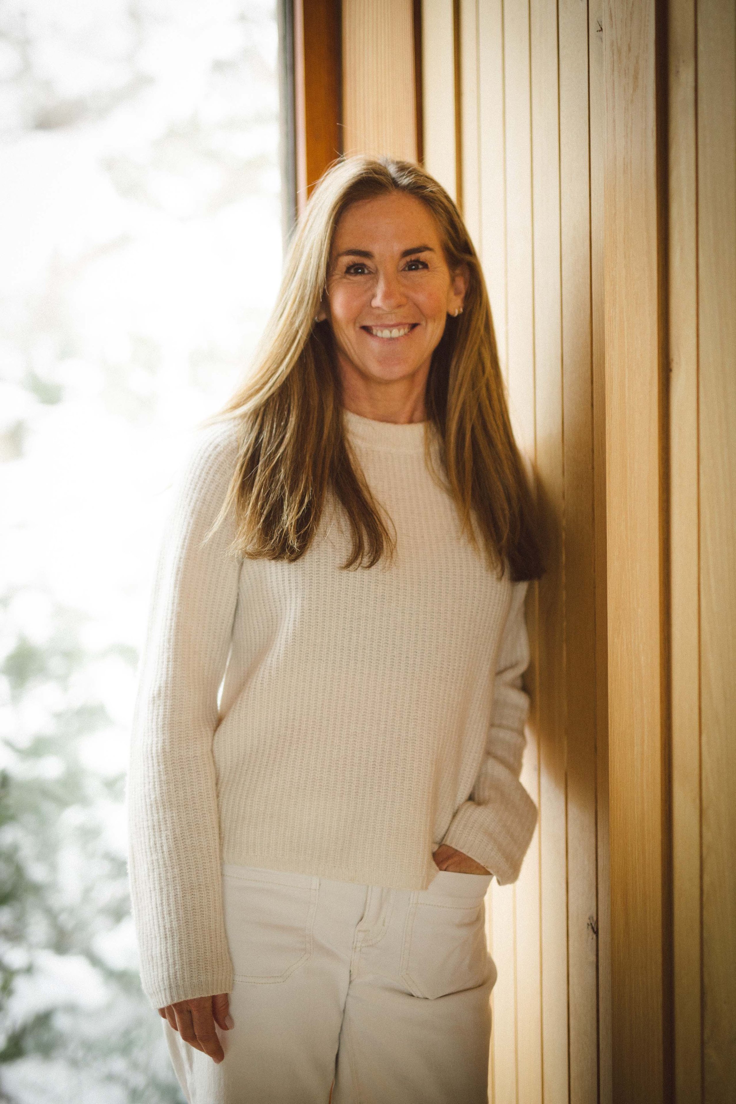 A woman with long, light brown hair smiling while standing indoors by a wooden wall, wearing a cream-colored sweater and light-colored pants.