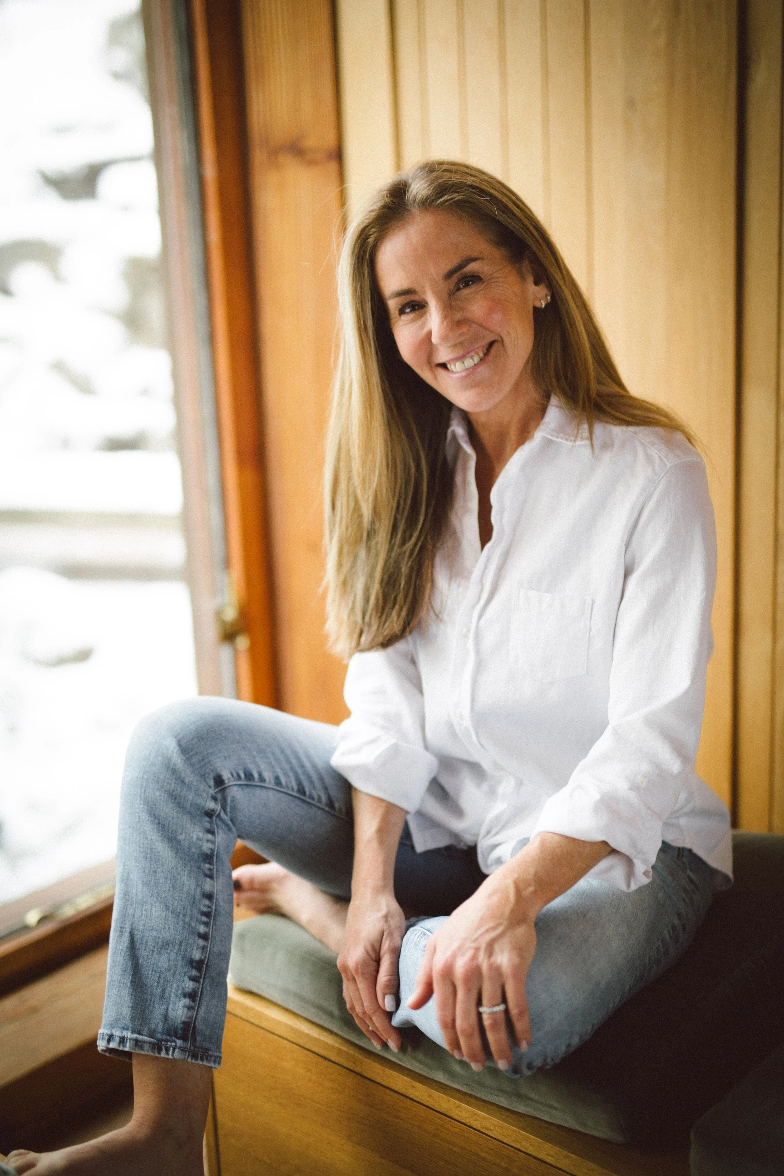 A woman with long brown hair, wearing a white button-up shirt and light blue jeans, sitting on a wooden bench by a window with snow outside, smiling at the camera.