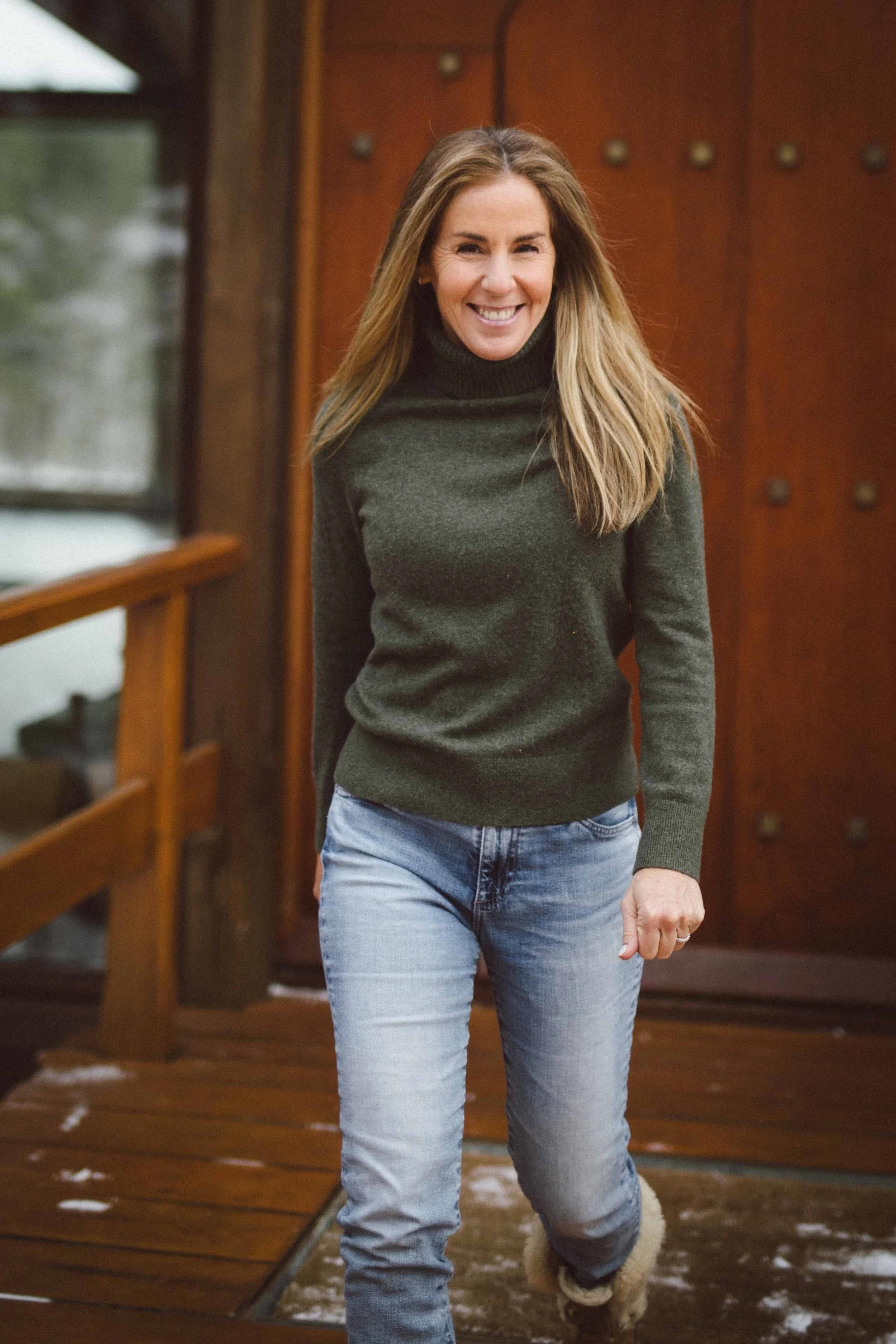 A woman smiling and walking on a wooden deck in winter, dressed in a dark green turtleneck sweater, light blue jeans, and furry boots.
