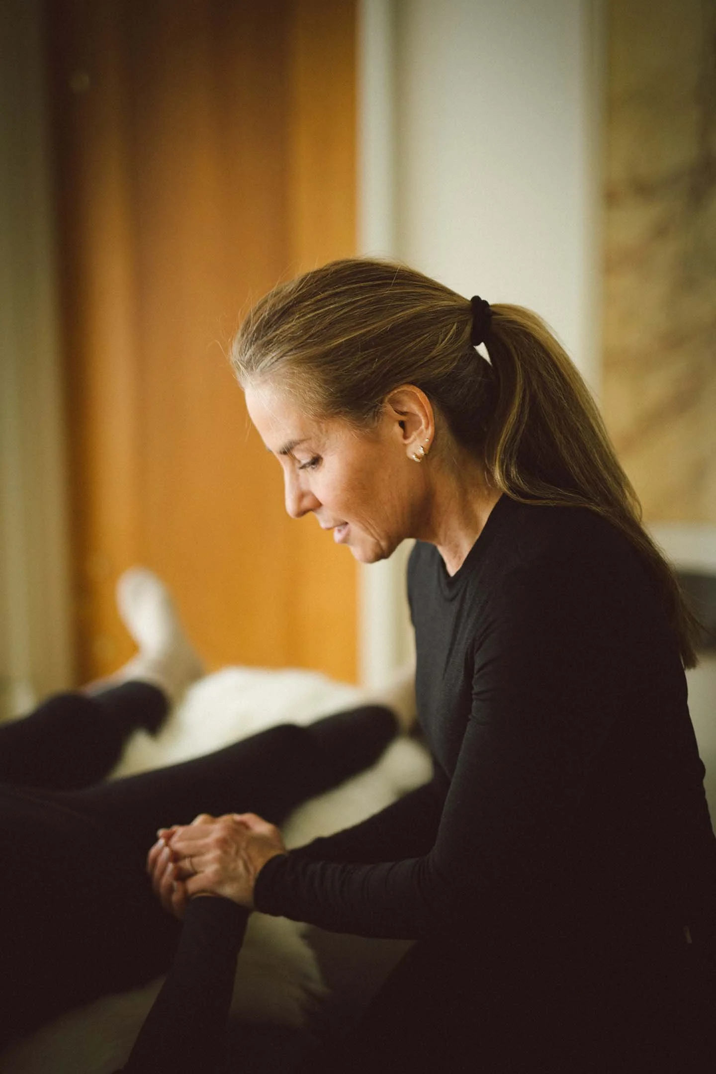 A woman with brown hair tied back, wearing a black long-sleeve shirt, is sitting with her head bowed and hands clasped in prayer or reflection in a warmly lit room.