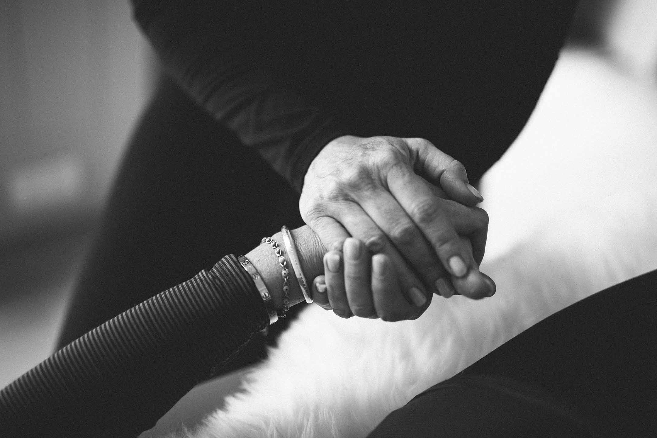 Black and white photo of two people holding hands, one with a thinner arm and wearing multiple bracelets. The other has a thicker arm and wears a dark long sleeve. The background is blurred.