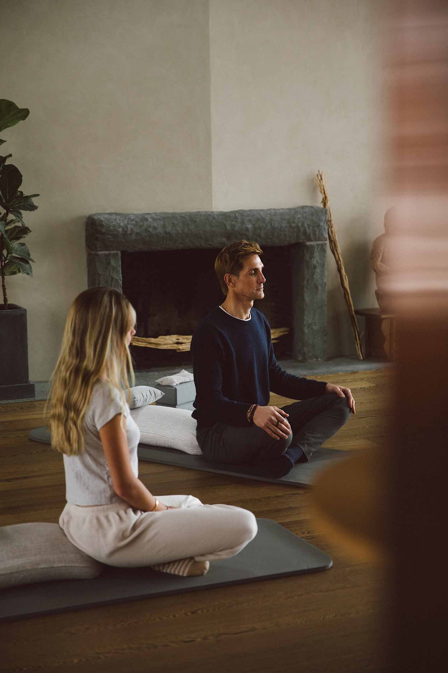 A man and a girl sitting cross-legged on yoga mats indoors, meditating in front of a fireplace with a large plant on the left and a decorative sculpture on a table on the right.