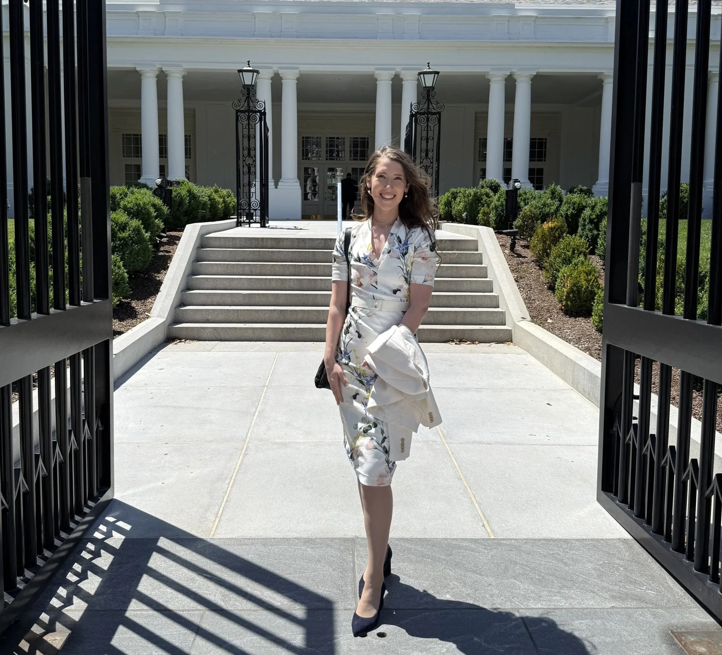 A woman walking into the White House, holding a white jacket and wearing a floral dress and blue heels.