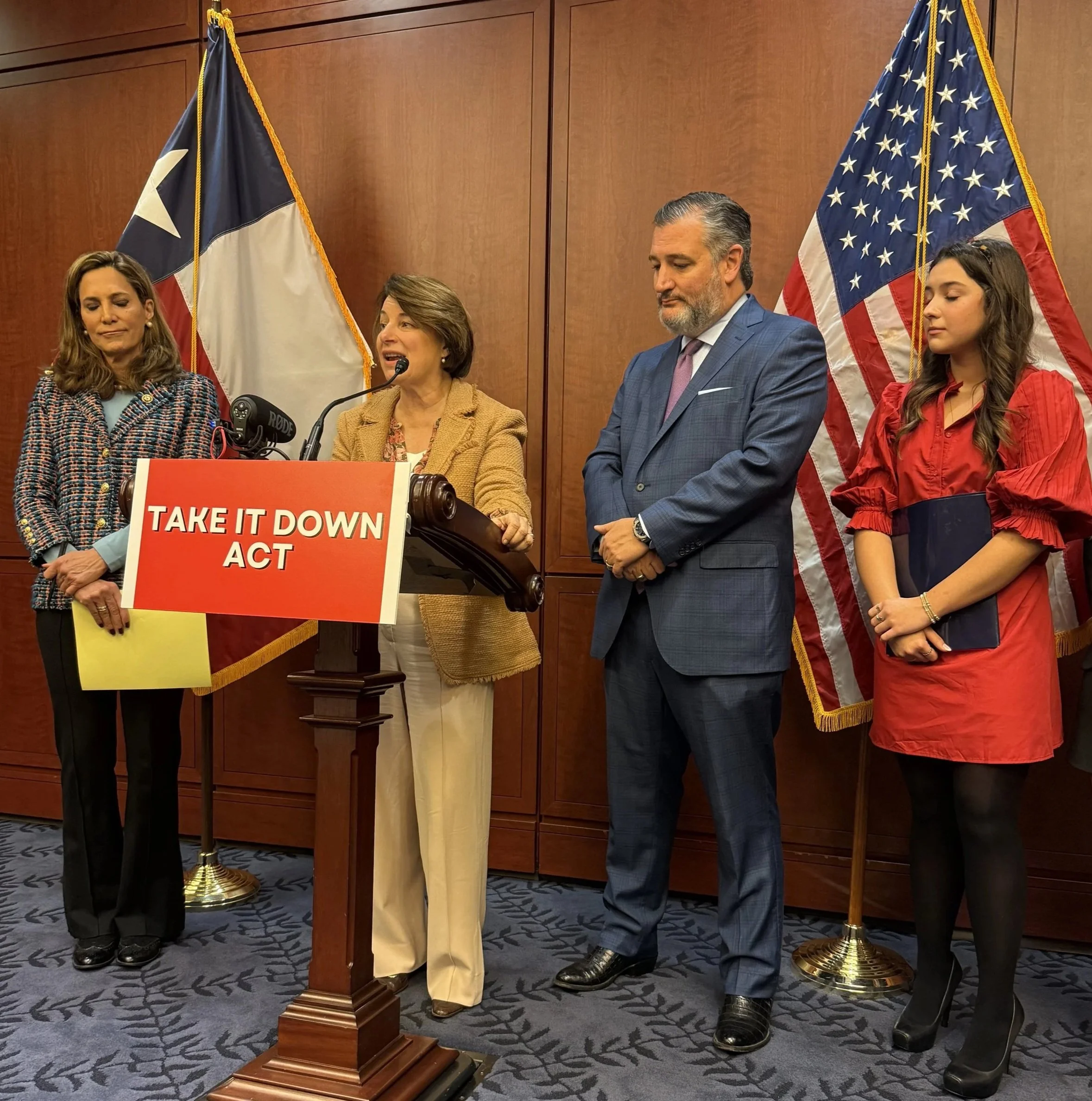 A group of four people standing in front of a wooden wall with two American flags. One woman is speaking at a podium that has a sign reading 'Take It Down Act'.