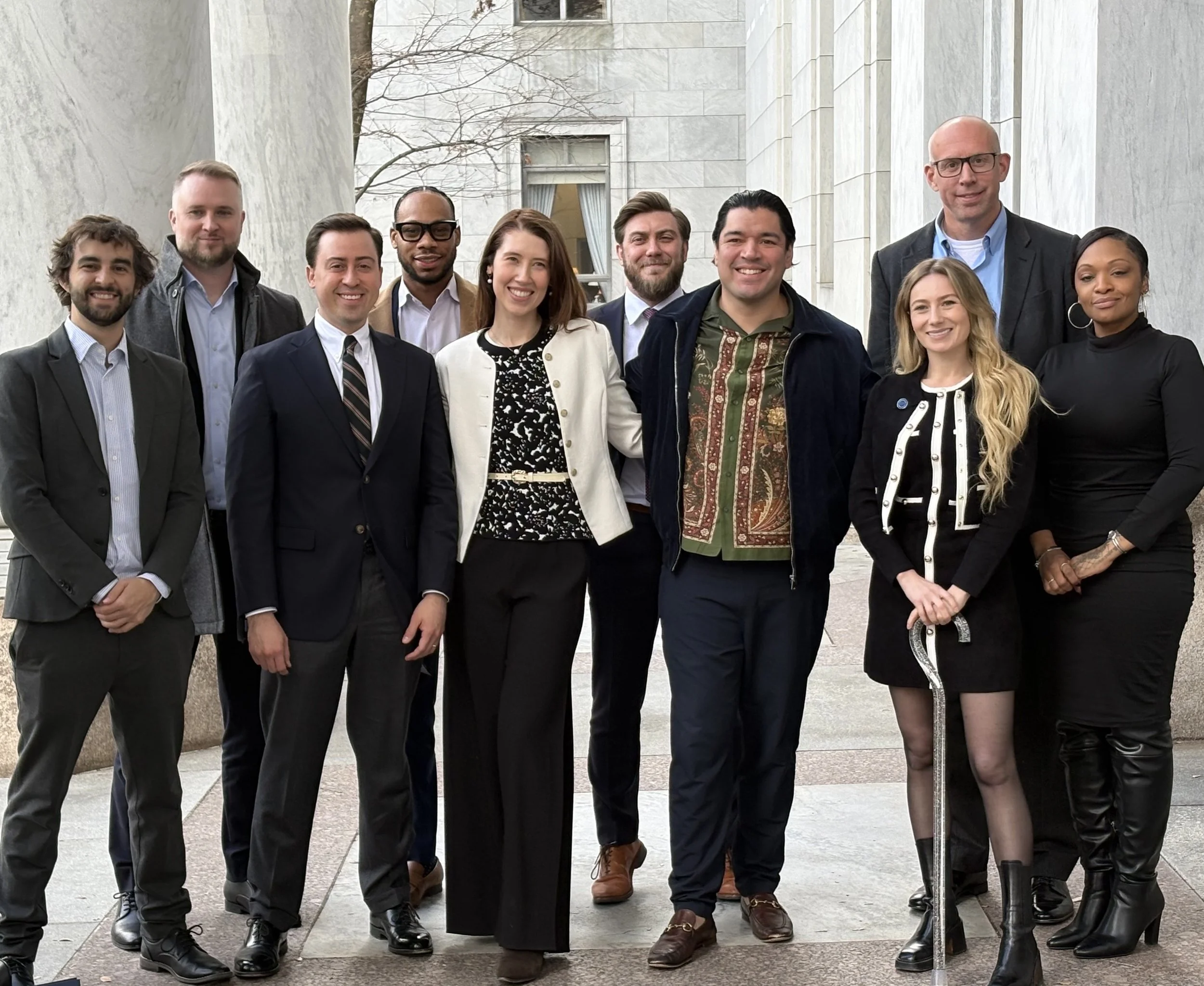 Group of twelve ethnically diverse professionally dressed people standing outdoors in front of a modern building, smiling at the camera.