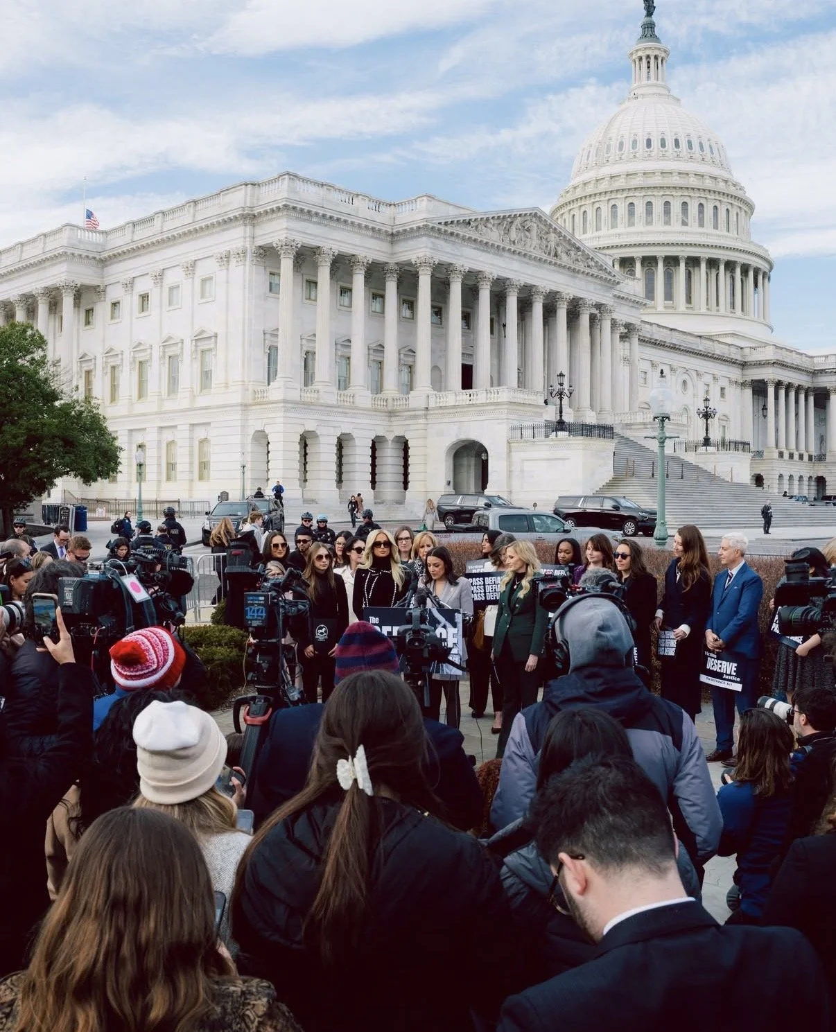A crowd gathers for a press conference outside the U.S. Capitol building in Washington D.C., with people holding signs and speaking into microphones.