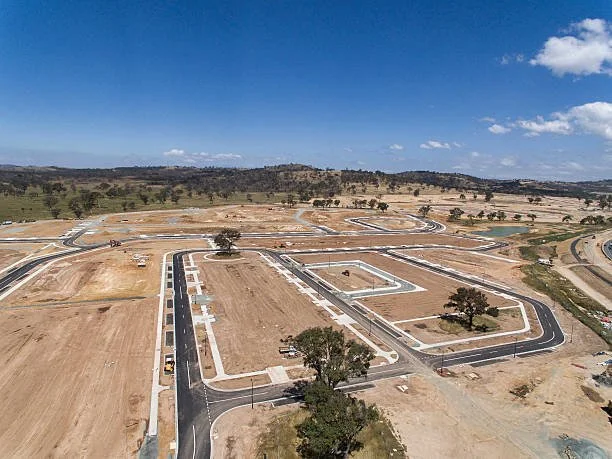 An aerial view of a new residential subdivision construction site in Perth WA, with newly paved roads, open land, and some scattered trees under a partly cloudy sky.