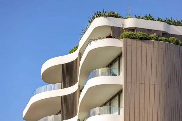 Modern apartment building with curvy white balconies, glass railings, and rooftop greenery against a clear blue sky.