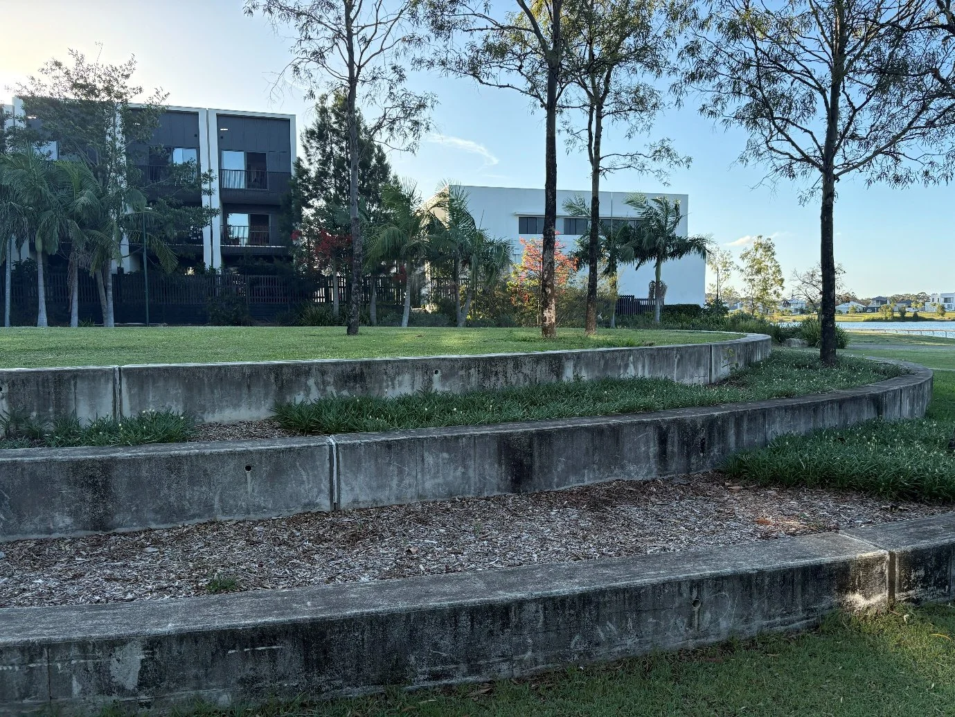 A multi-tiered garden with concrete retaining walls, green grass, and trees in the foreground, with modern residential buildings and a water body in the background.