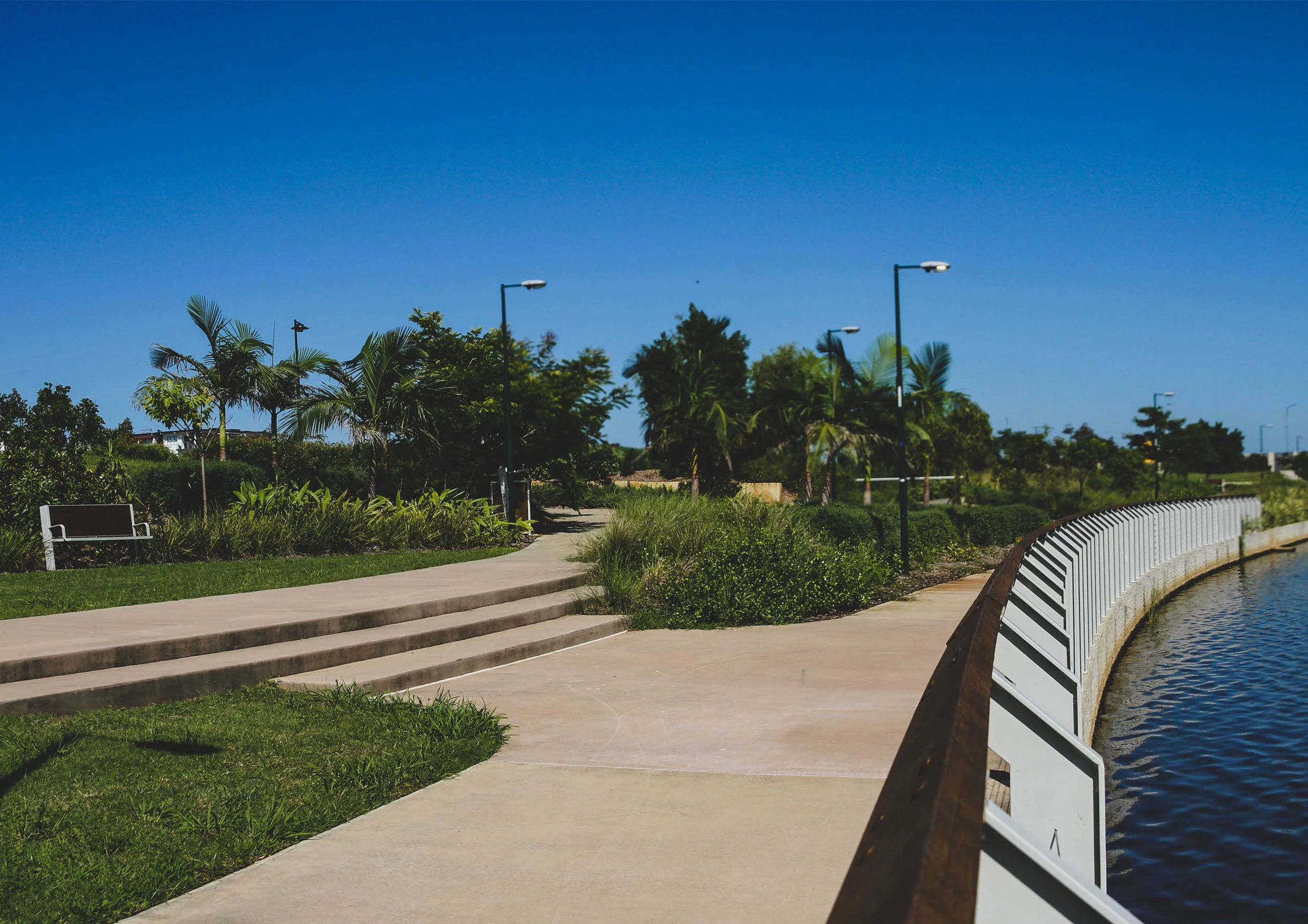 A peaceful park scene with a concrete pathway, green grass, and bushes on the left side. Tall palm trees and other trees are visible in the background under a clear blue sky. There are streetlights along the pathway, and a white fence curves along a body of water on the right side.