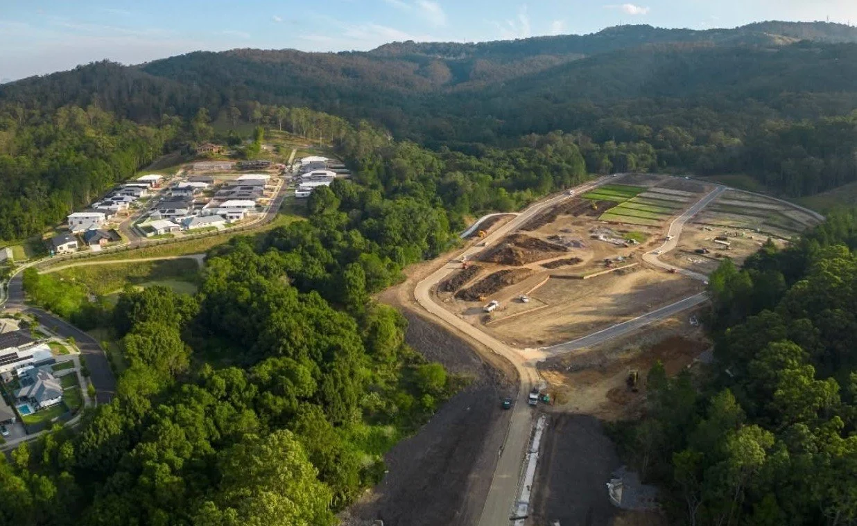 Aerial view of a multistage hillside subdivision with houses and a construction site in progress, surrounded by lush green trees and distant mountains.