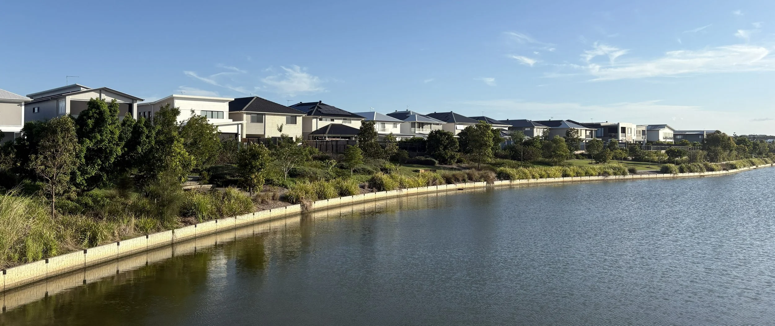 Modern houses lining a waterway with trees and shrubs along the shore, under a clear blue sky.