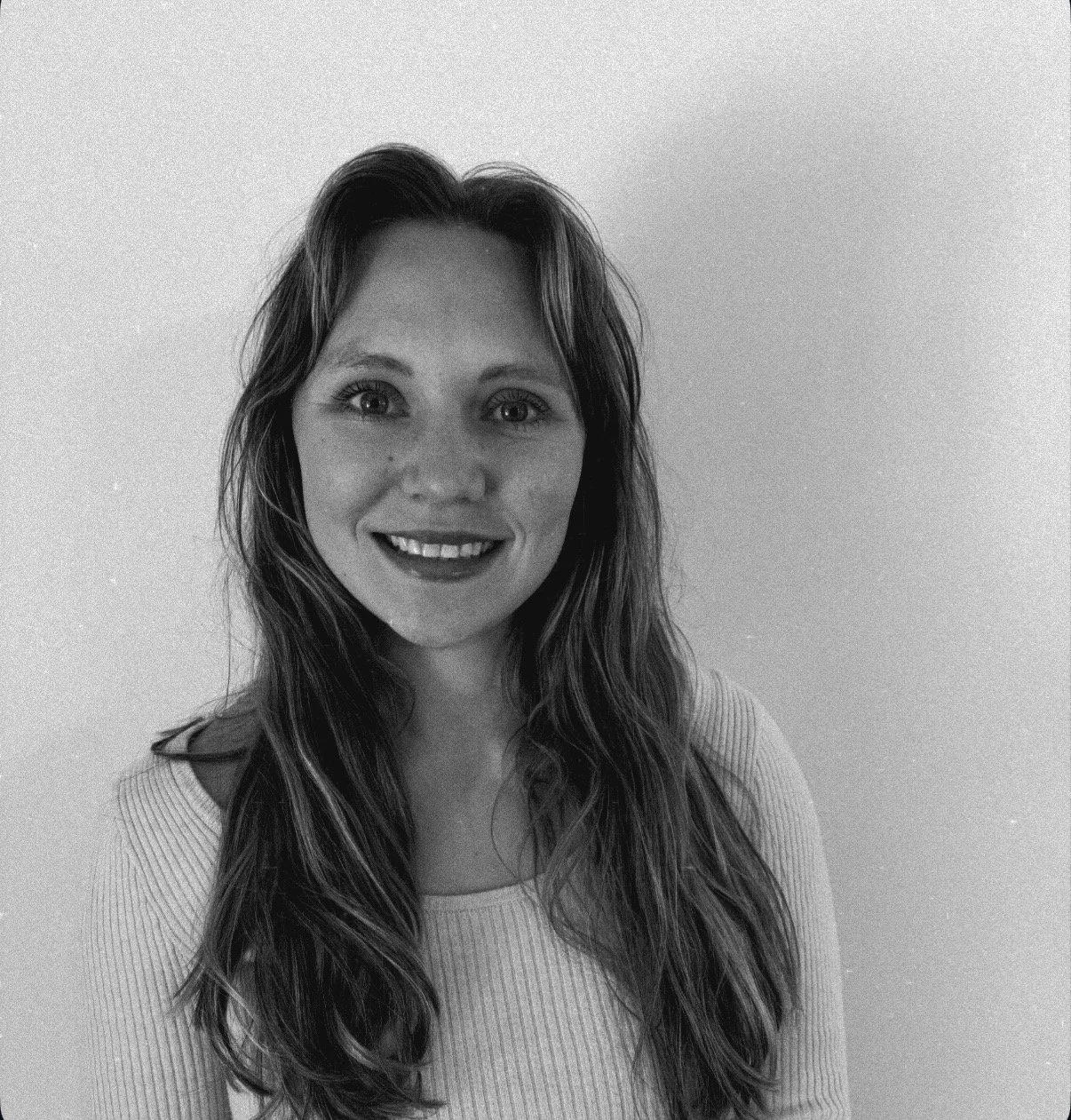 Young woman with long wavy hair smiling, wearing a light-colored top, against a plain background.