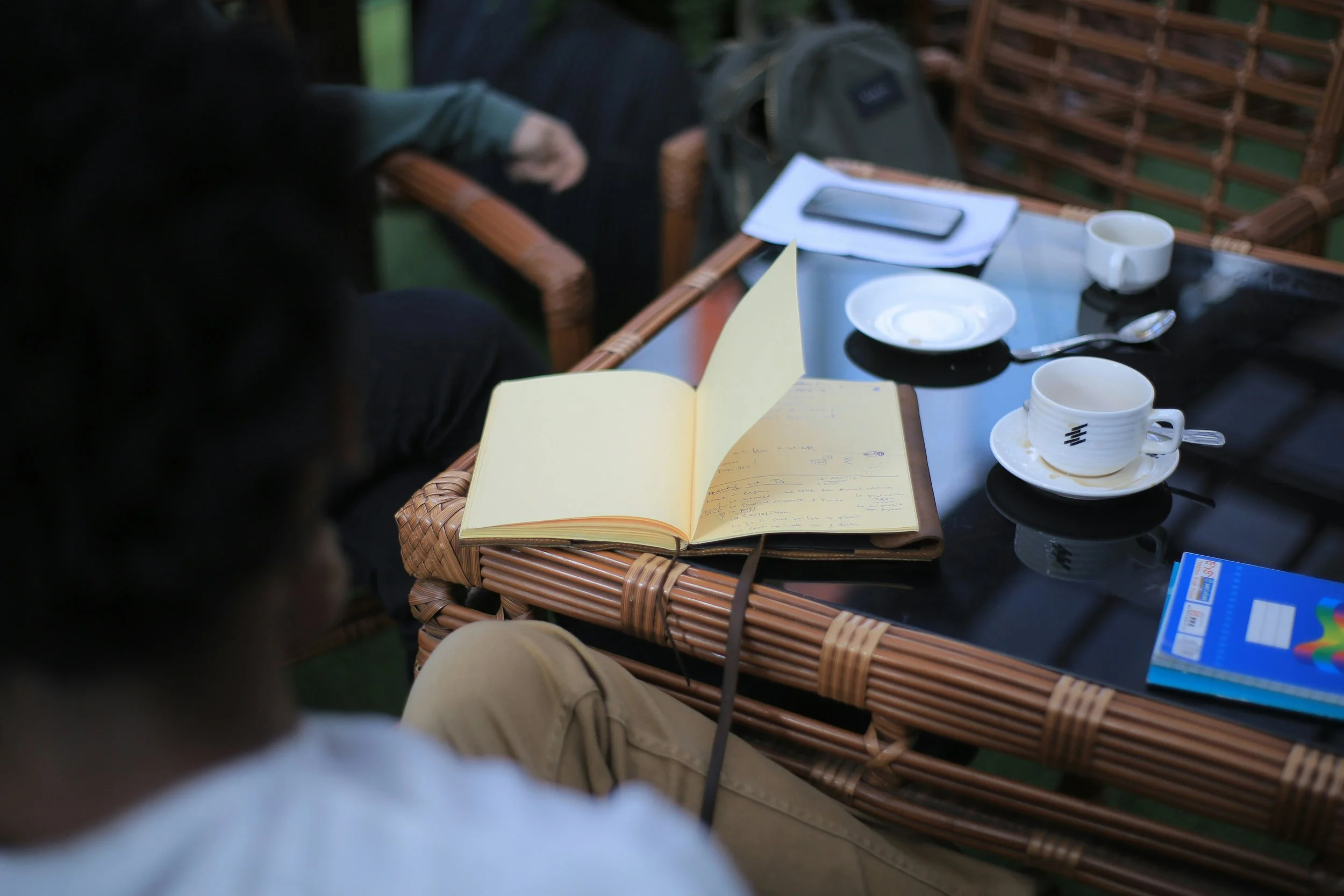 A table outdoors with an open notebook, coffee cups, a spoon, a smartphone, a notebook, and some paper on a glass-top wicker table, with some people sitting around it.