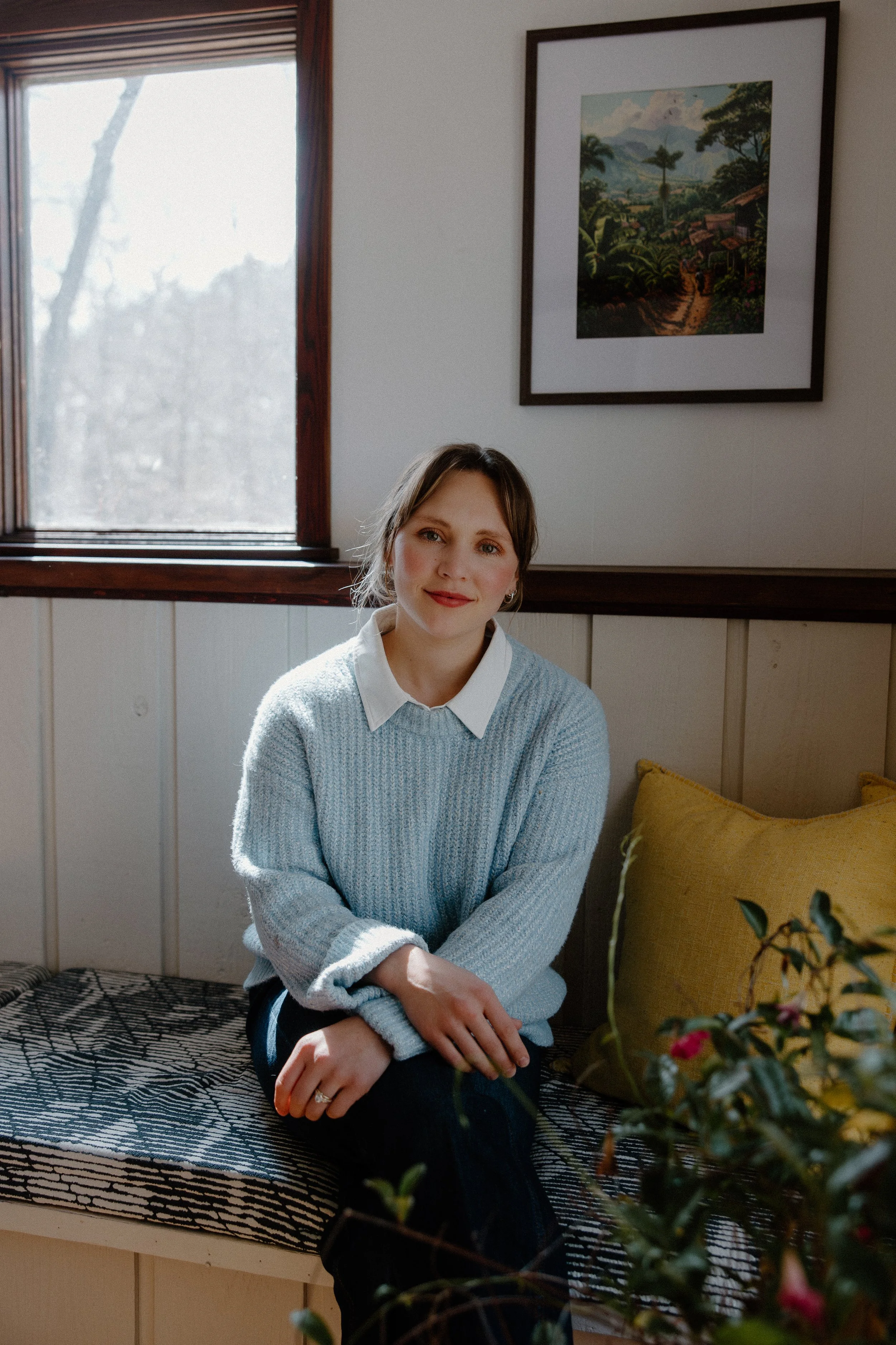 A young woman with shoulder-length brown hair and earrings, sitting on a patterned bench in a room with a window and framed artwork on the wall. She is wearing a light blue sweater over a white collared shirt, with her hands resting on her lap and smiling slightly at the camera. There is a yellow cushion beside her and some plants in the foreground.