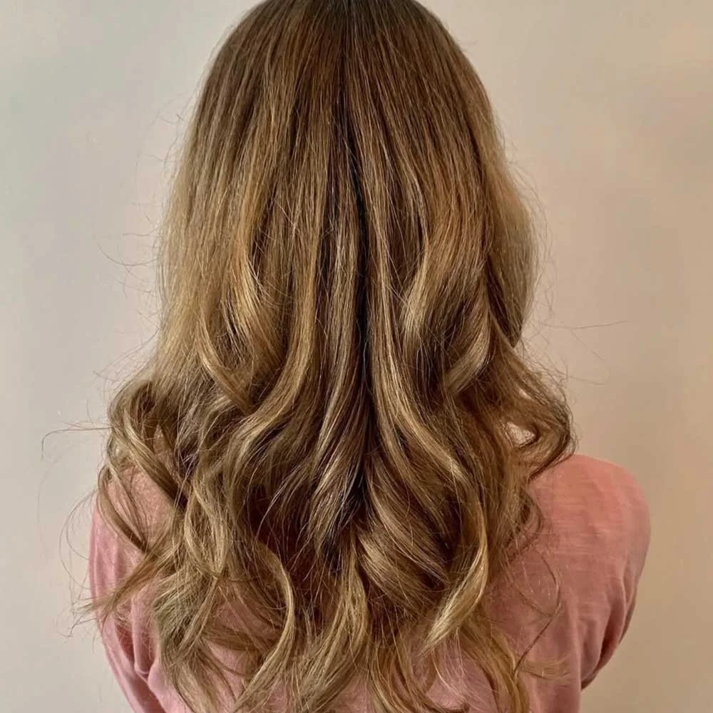 Back view of a woman with long, wavy, caramel-colored hair wearing a pink top.