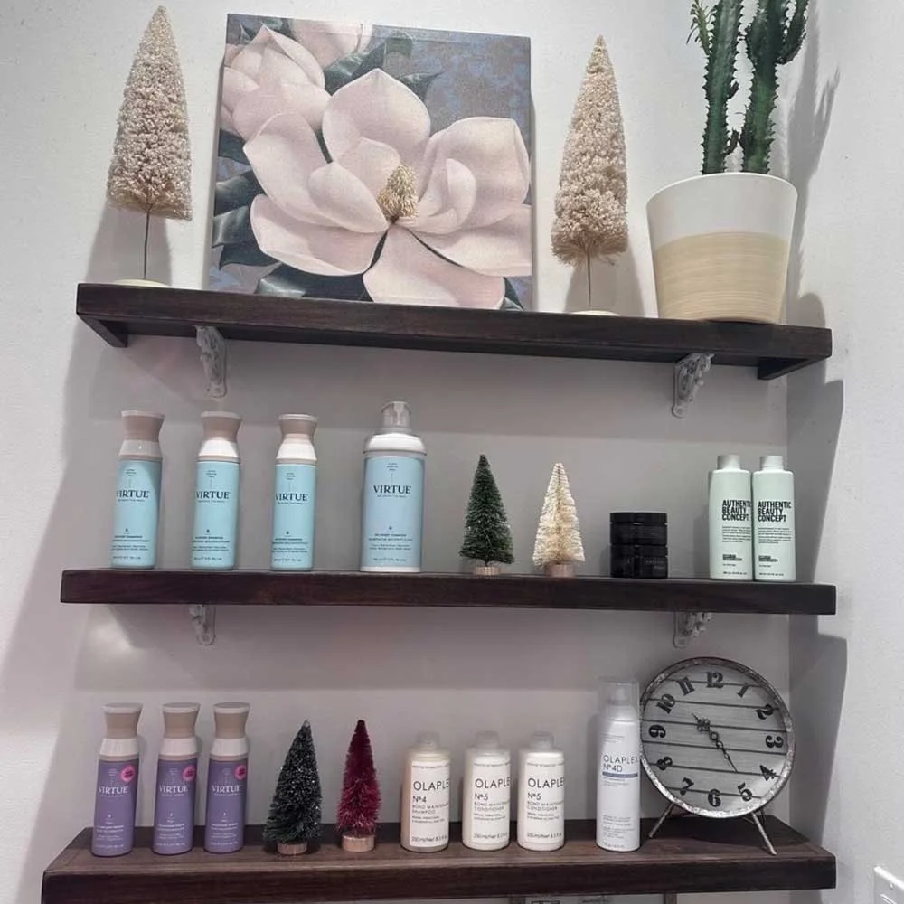 Decorative wall shelves with haircare, skincare, and cosmetic bottles, small artificial Christmas trees, a clock, and potted plants, with artwork of a flower above.