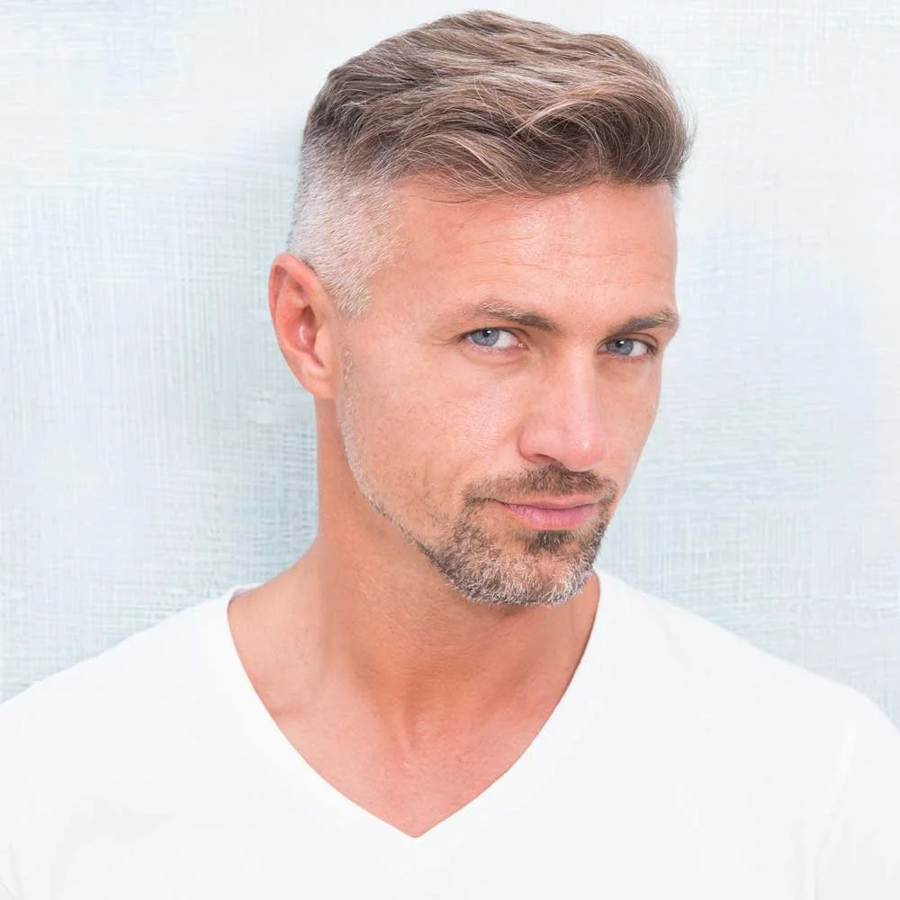 Close-up of a man with styled blonde hair, light stubble, and blue eyes wearing a white shirt, with a soft-focus light-colored background.