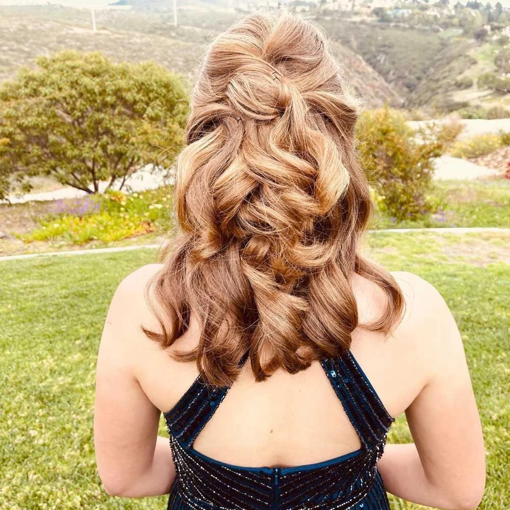 A woman with long, wavy, light brown hair standing outdoors in a grassy area with trees and hills in the background