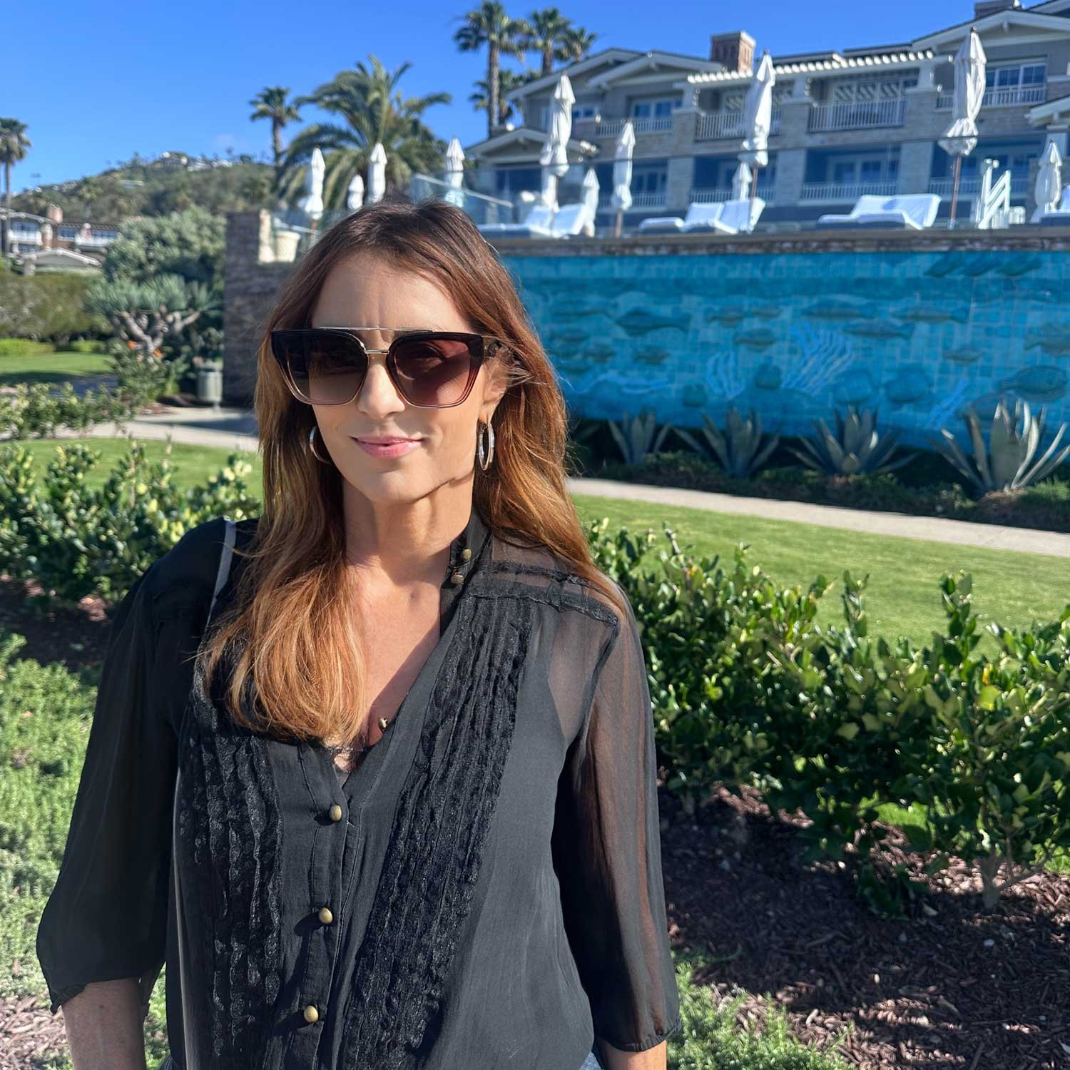 A woman with long brown hair, wearing large sunglasses, a black sheer blouse, and hoop earrings, standing outdoors in front of a landscaped garden with a swimming pool and a large house in the background on a sunny day.