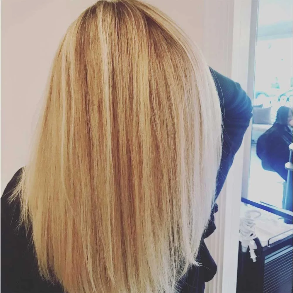 Woman with long, straight blonde hair leaning forward inside a salon or similar setting.