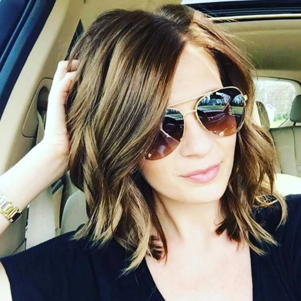 A woman with short, wavy brown hair wearing sunglasses and a black top, sitting inside a vehicle.
