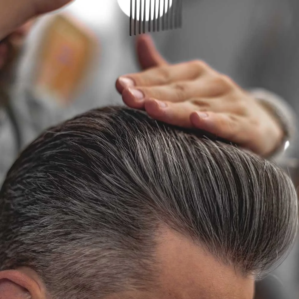 A person with middle gray hair is receiving a professional hair treatment or styling, with a hand brushing the hair and a comb held above the head.