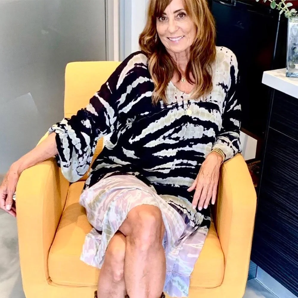 A woman with long red hair sitting on a yellow chair, smiling, wearing a black and white patterned dress.
