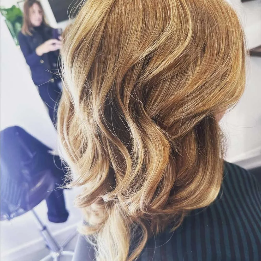 Close-up of a woman's wavy, blonde hair with highlights, sitting in a salon chair.