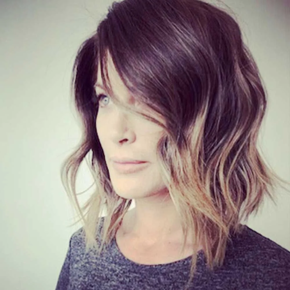 A woman with wavy, shoulder-length hair that is dark brown with lighter brown highlights, looking to her left with a slight smile, wearing a dark shirt against a plain background.