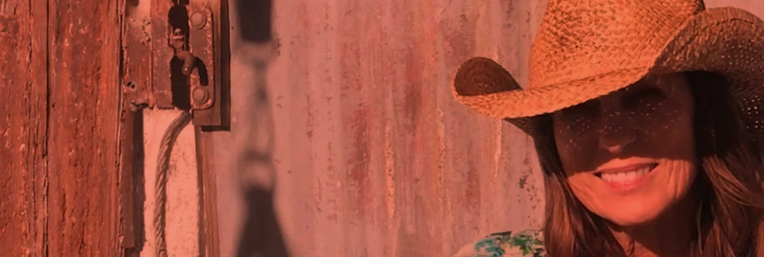 A woman wearing a wide-brimmed woven hat and sunglasses, smiling, standing near a chain-link fence at sunset or sunrise, with a wooden fence and outdoor setting in the background.