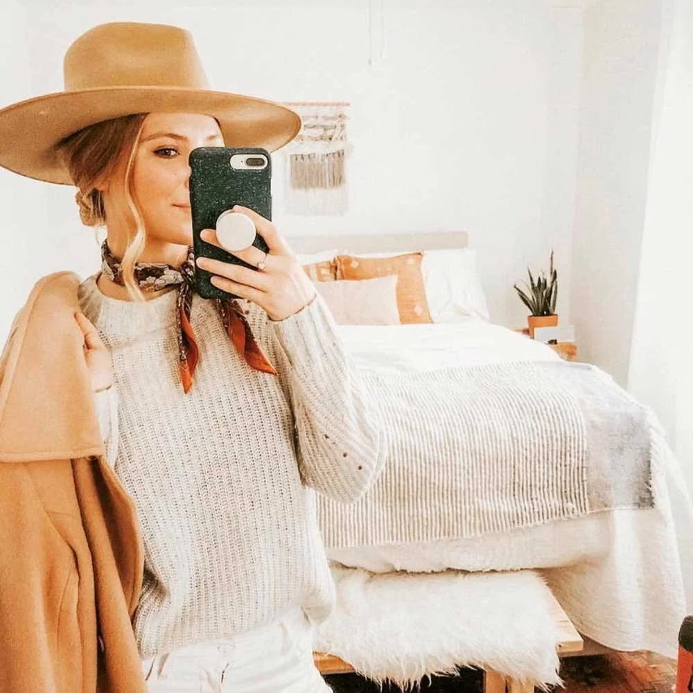 Woman taking a mirror selfie in a bedroom. She is wearing a large tan wide-brim hat, a beige sweater, a scarf around her neck, and has light brown hair styled in waves.